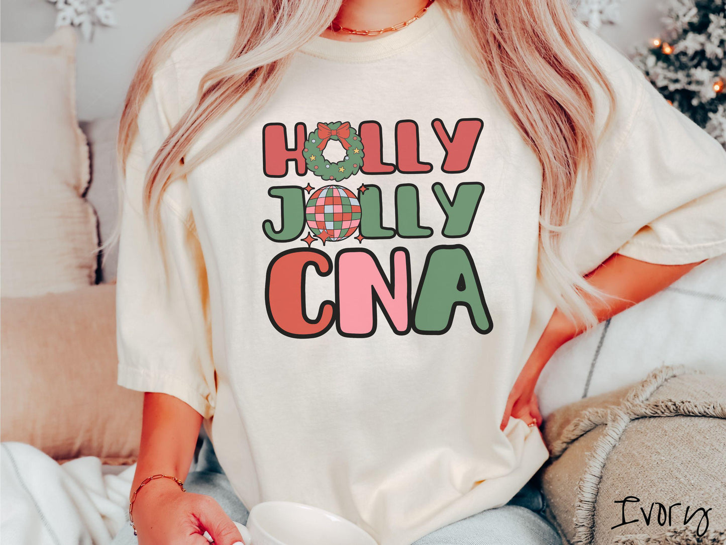 A woman wearing a cute, vintage ivory colored Comfort Colors T-shirt with the text Holly Jolly CNA in red, green, pink, and blue font. The letter O in Holly is a Christmas wreath and the letter O in Jolly is a multi-colored disco ball.