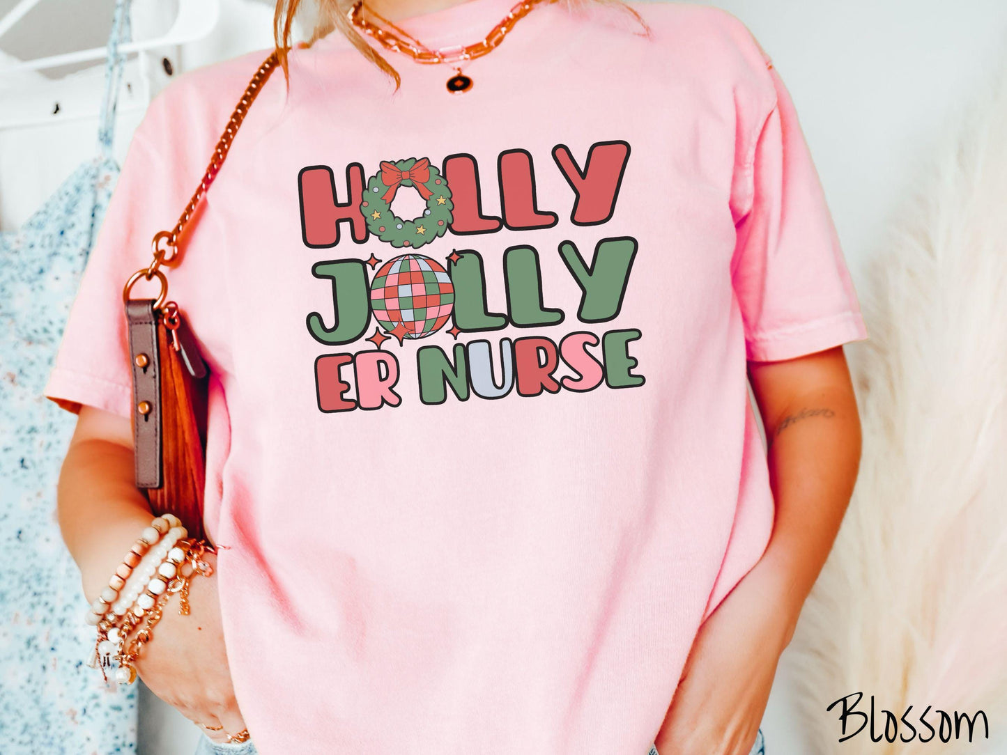 A woman wearing a cute, vintage blossom colored Comfort Colors T-shirt with the text Holly Jolly ER Nurse in red, green, pink, and blue font. The letter O in Holly is a Christmas wreath and the letter O in Jolly is a multi-colored disco ball.