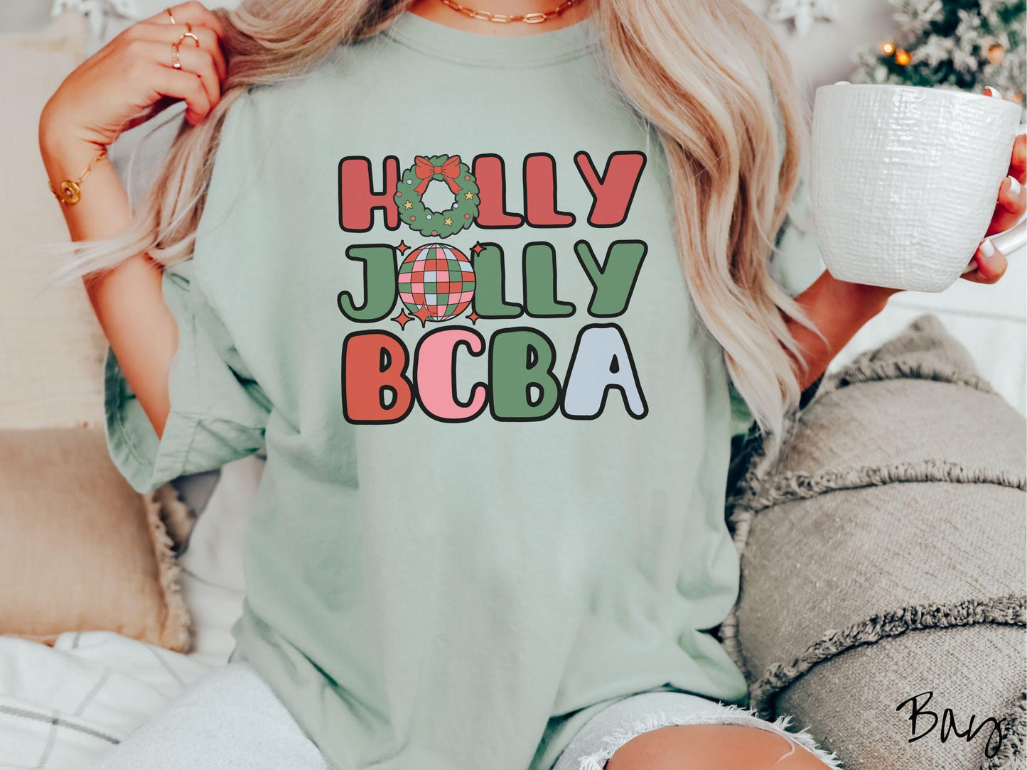 A woman wearing a cute, vintage bay colored Comfort Colors T-shirt with the text Holly Jolly BCBA in red, green, pink, and blue font. The letter O in Holly is a Christmas wreath and the letter O in Jolly is a multi-colored disco ball.