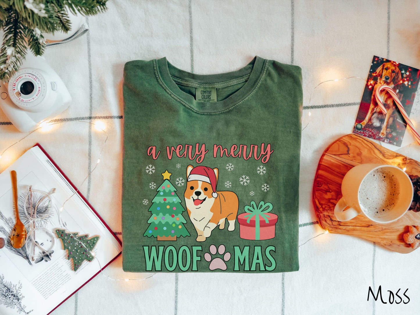 A vintage moss colored Comfort Colors T-shirt with the text 'a very merry Woofmas' in red and green font, featuring an orange corgi wearing a red Santa hat sitting between a Christmas tree and a present with snowflakes hanging in the air.