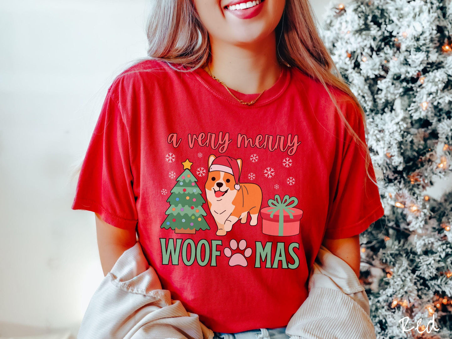 A vintage red colored Comfort Colors T-shirt with the text 'a very merry Woofmas' in red and green font, featuring an orange corgi wearing a red Santa hat sitting between a Christmas tree and a present with snowflakes hanging in the air.