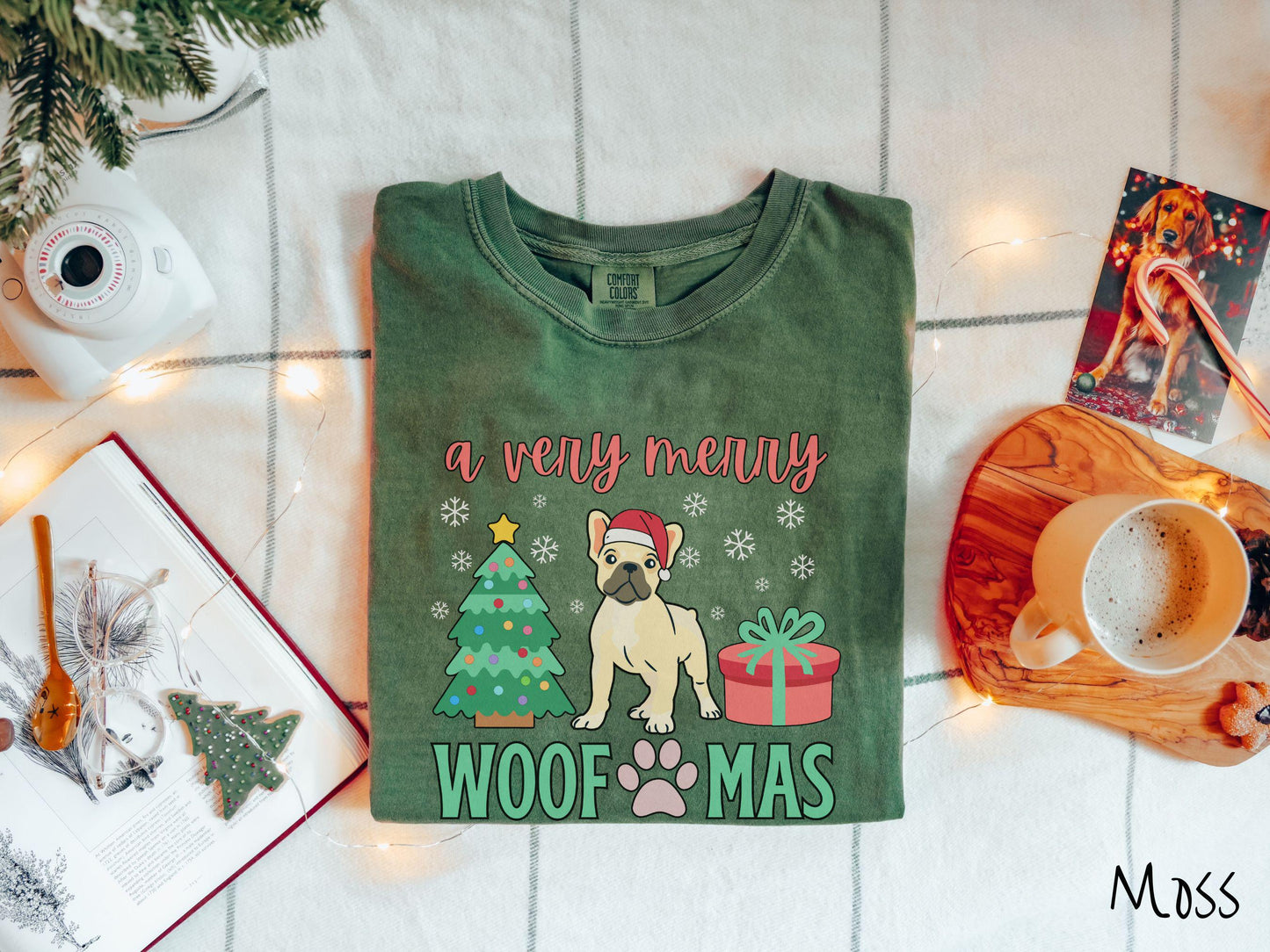 A vintage moss colored Comfort Colors T-shirt with the text 'a very merry Woofmas' in red and green font, featuring a French Bulldog wearing a red Santa hat sitting between a Christmas tree and a present with white snowflakes hanging in the air.