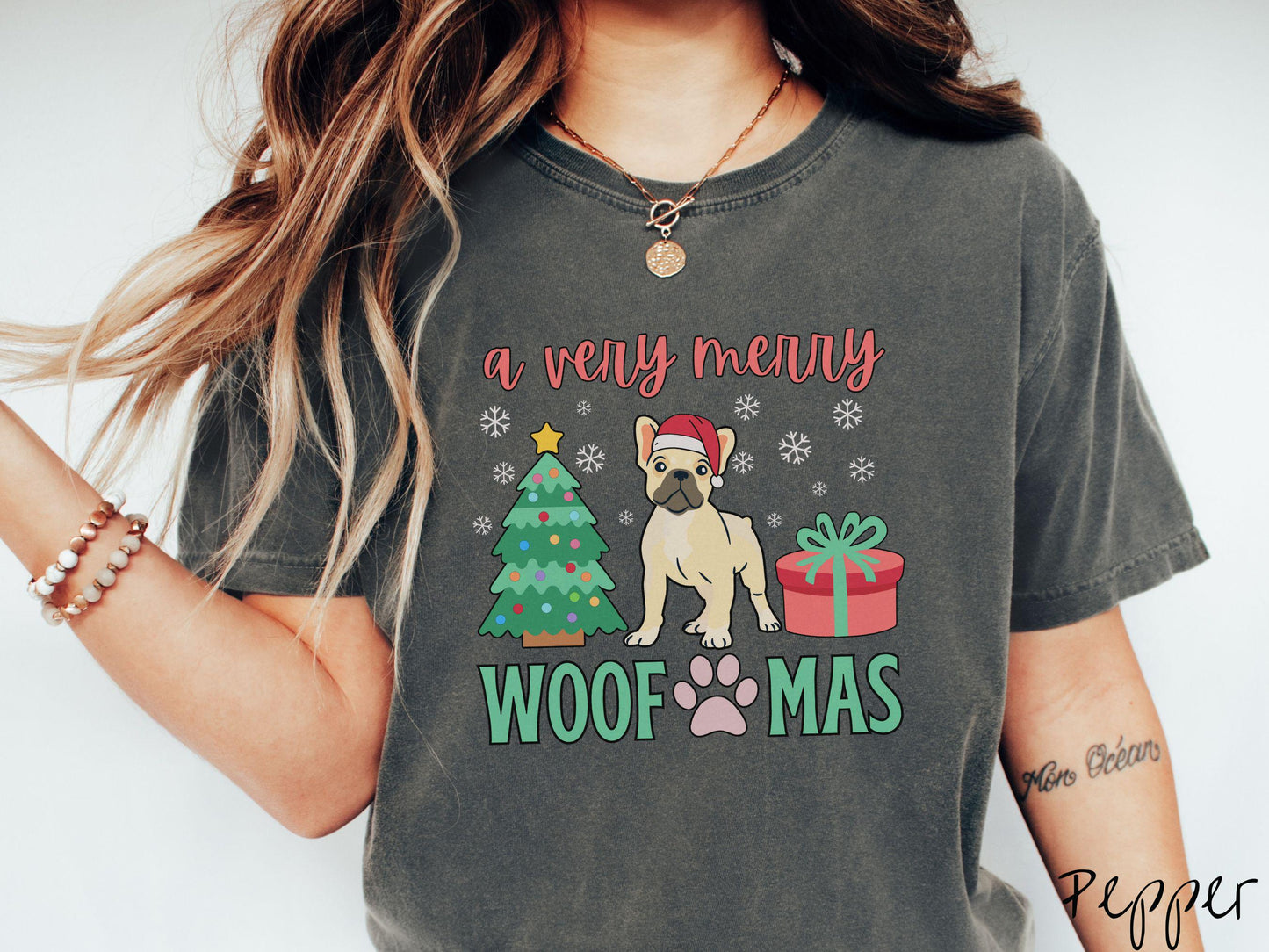 A vintage pepper colored Comfort Colors T-shirt with the text 'a very merry Woofmas' in red and green font, featuring a French Bulldog wearing a red Santa hat sitting between a Christmas tree and a present with white snowflakes hanging in the air.