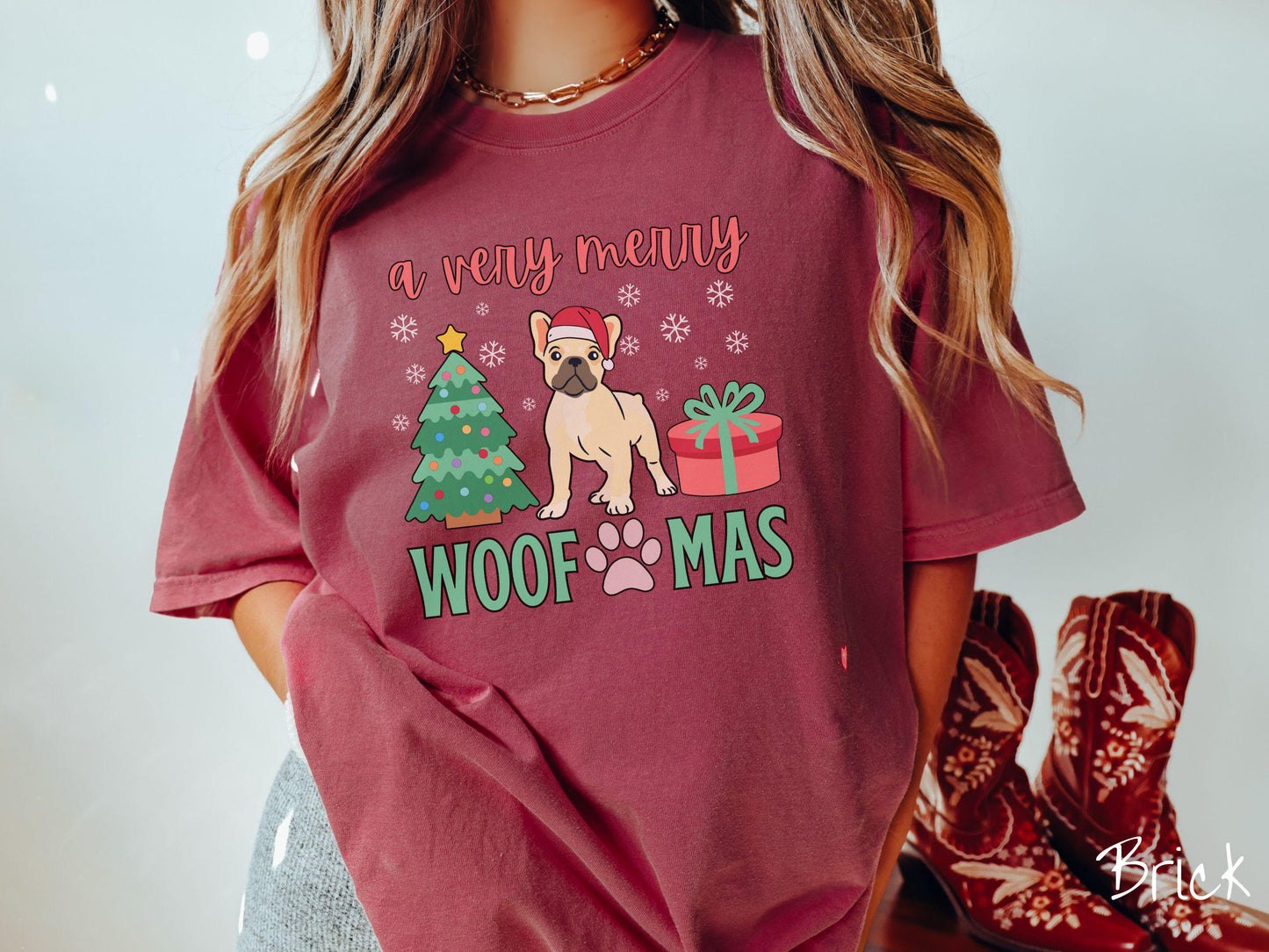 A vintage brick colored Comfort Colors T-shirt with the text 'a very merry Woofmas' in red and green font, featuring a French Bulldog wearing a red Santa hat sitting between a Christmas tree and a present with white snowflakes hanging in the air.