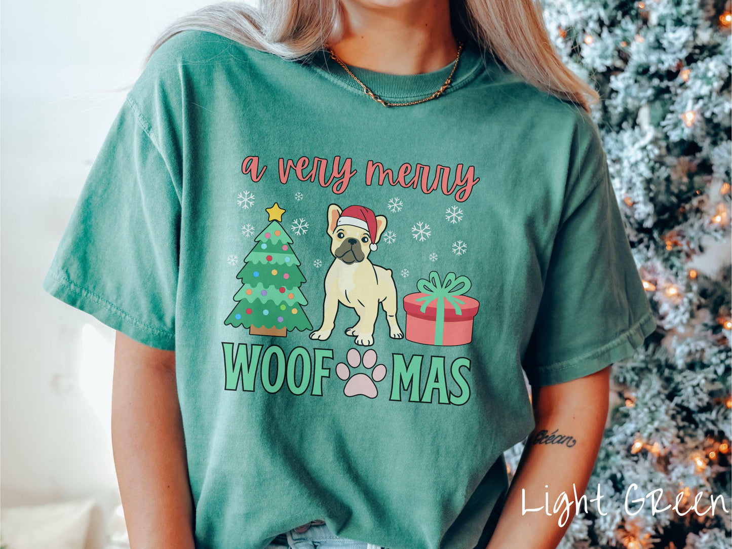 A vintage light green colored Comfort Colors T-shirt with the text 'a very merry Woofmas' in red and green font, featuring a French Bulldog wearing a red Santa hat sitting between a Christmas tree and a present with snowflakes hanging in the air.
