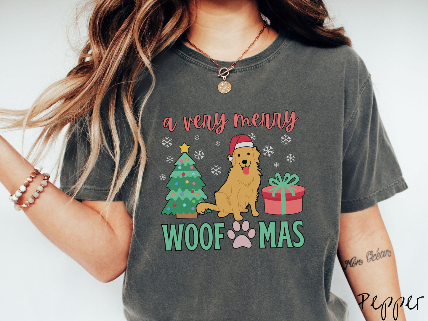 A vintage pepper colored Comfort Colors T-shirt with the text 'a very merry Woofmas' in red and green font, featuring a Golden Retriever wearing a red Santa hat sitting between a Christmas tree and a present with snowflakes hanging in the air.