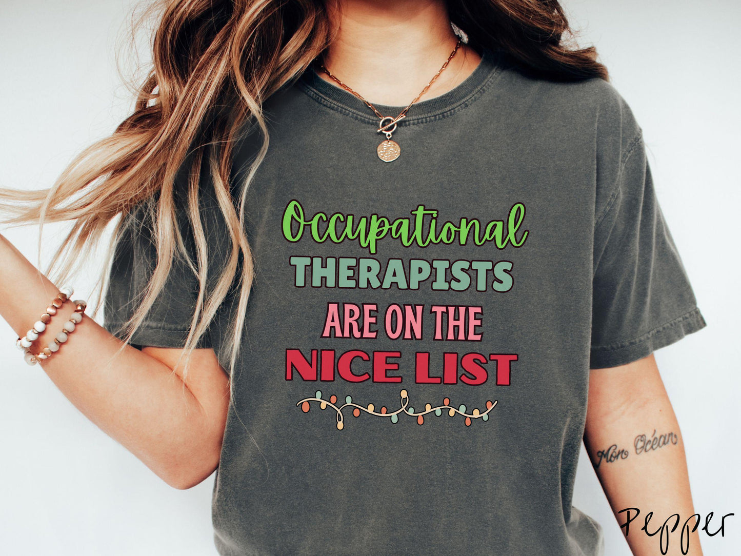 A vintage pepper colored Comfort Colors T-shirt features the text 'Occupational Therapy is on the nice list' in green, pink, and red fonts. There is a string of colorful Christmas lights along the bottom, exuding a cozy holiday vibe.