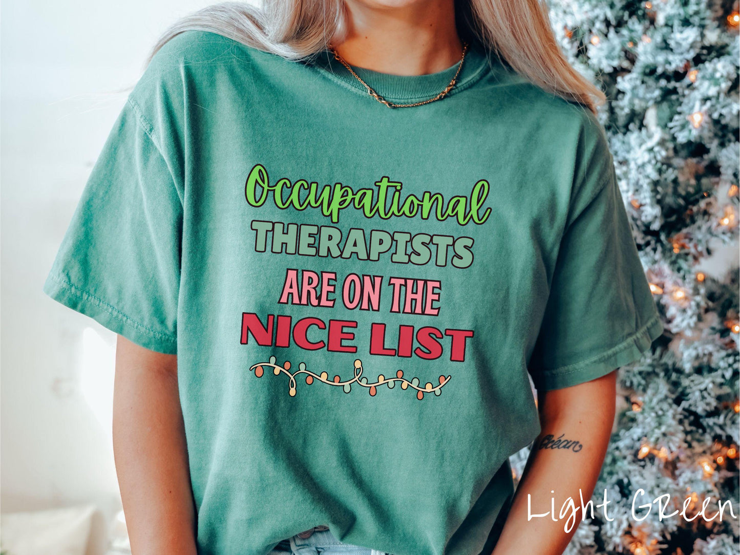 A vintage light green colored Comfort Colors T-shirt features the text 'Occupational Therapy is on the nice list' in green, pink, and red fonts. There is a string of colorful Christmas lights along the bottom, exuding a cozy holiday vibe.