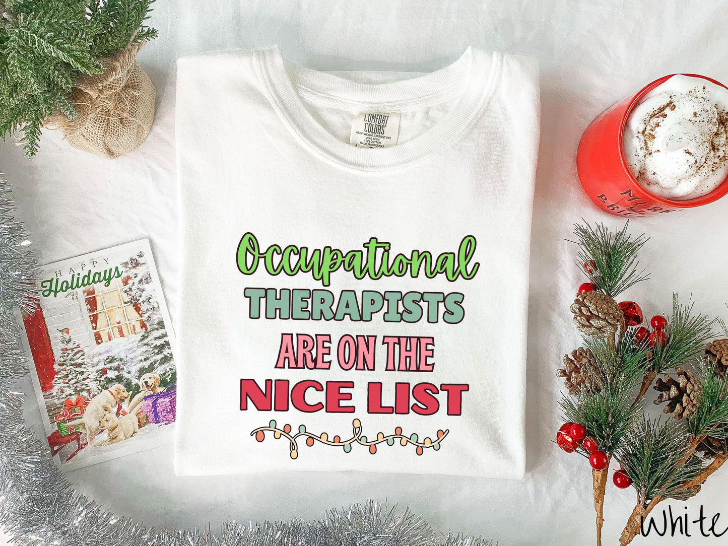 A vintage white colored Comfort Colors T-shirt features the text 'Occupational Therapy is on the nice list' in green, pink, and red fonts. There is a string of colorful Christmas lights along the bottom, exuding a cozy holiday vibe.
