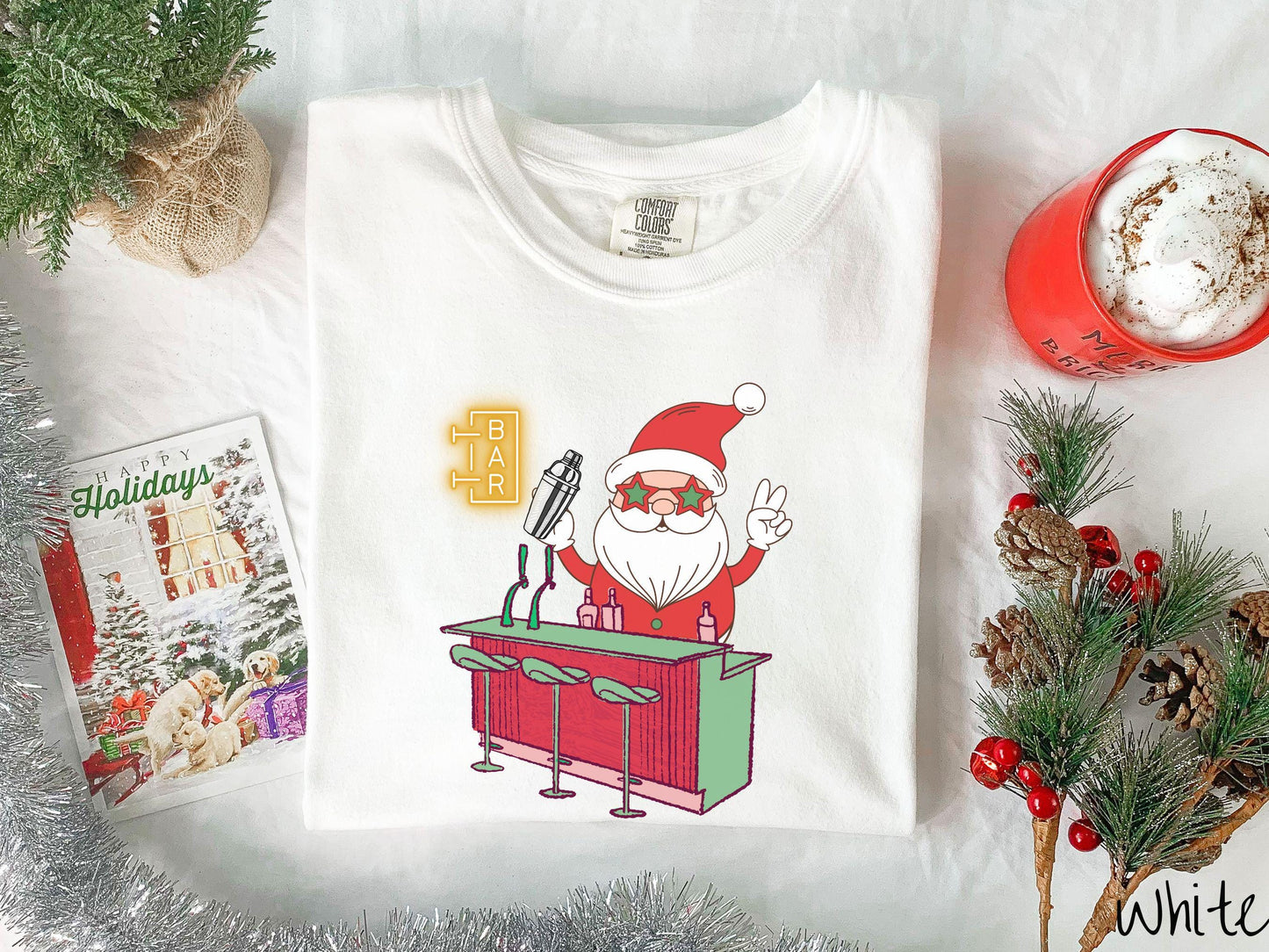 A vintage white colored Comfort Colors T-shirt features Santa Claus with red and green glasses mixing drinks and holding up a peace sign behind a red and green bar.