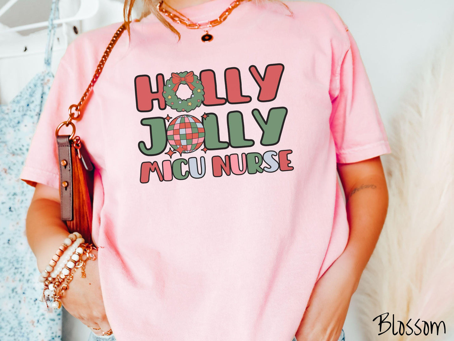 A woman wearing a cute, vintage blossom colored Comfort Colors T-shirt with the text Holly Jolly MICU Nurse in red, green, pink, and blue font. The letter O in Holly is a Christmas wreath and the letter O in Jolly is a multi-colored disco ball.