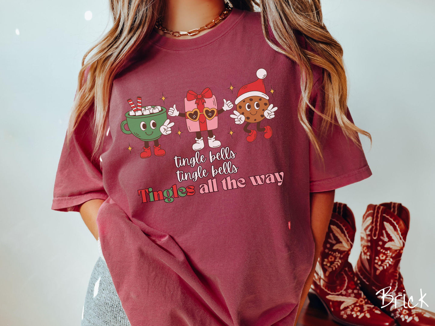 A cute, vintage brick colored Comfort Colors T-shirt with the text tnigle bells, tingle bells, tingles all the way. Above are a green cup of hot cocoa holding a peace sign, a red present holding two thumbs up, and a cookie holding up peace signs.