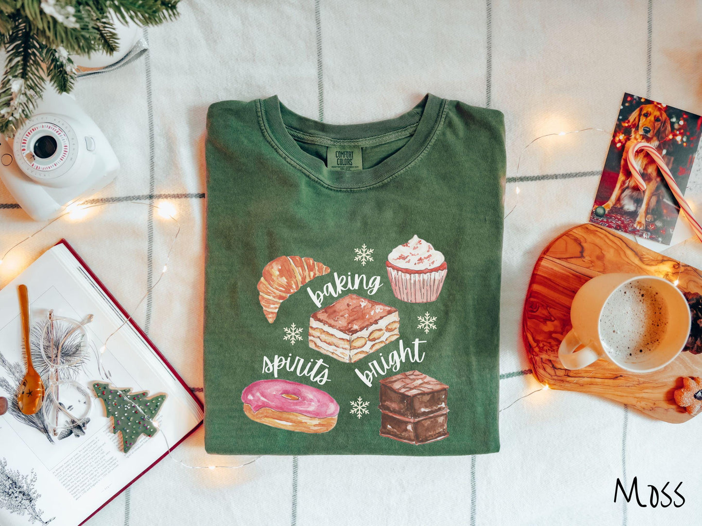 This vintage moss colored Comfort Colors T-shirt features the text 'baking spirits bright' in white cursive font, accompanied by cute dessert illustrations and white snowflakes.