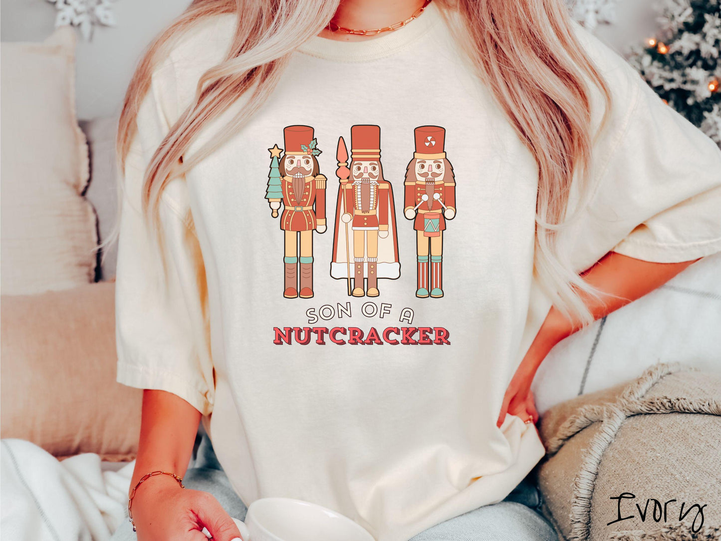 This vintage ivory colored Comfort Colors T-shirt features three red nutcracker figurines dressed in Christmas attire and one cracking a nut. Below is the text 'Son of a nutcracker' in red and white font.