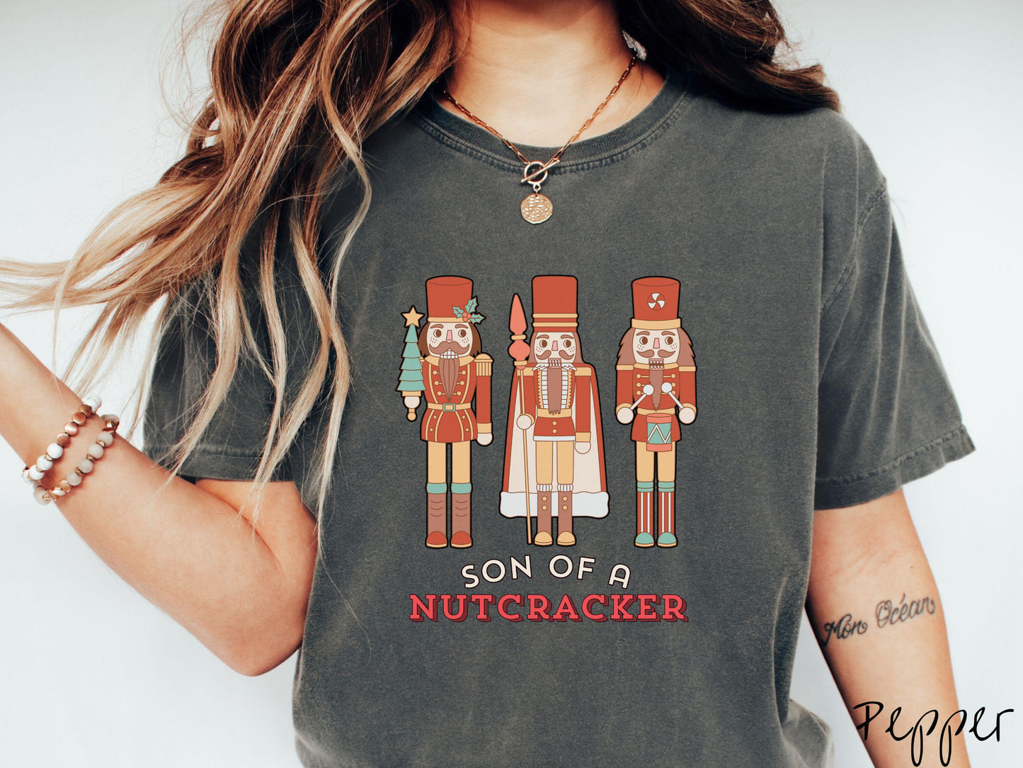 This vintage pepper colored Comfort Colors T-shirt features three red nutcracker figurines dressed in Christmas attire and one cracking a nut. Below is the text 'Son of a nutcracker' in red and white font.