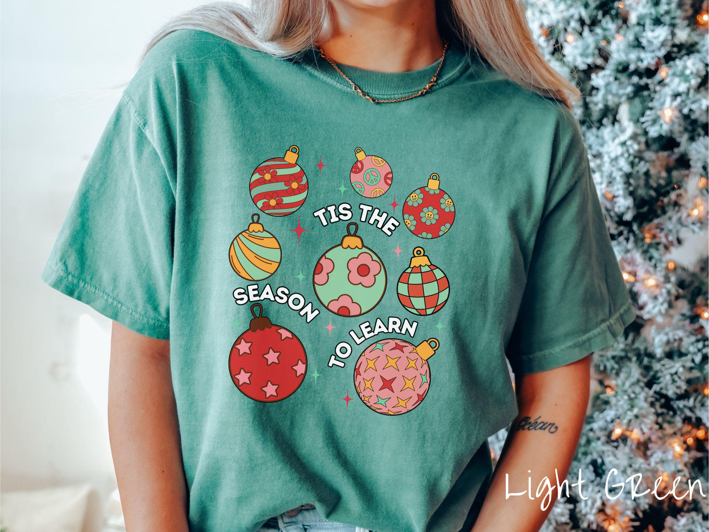 This vintage light green colored Comfort Colors T-shirt features eight colorful Christmas tree ornaments arranged in a grid pattern and twinkling stars with the text Tis the Season to Learn.
