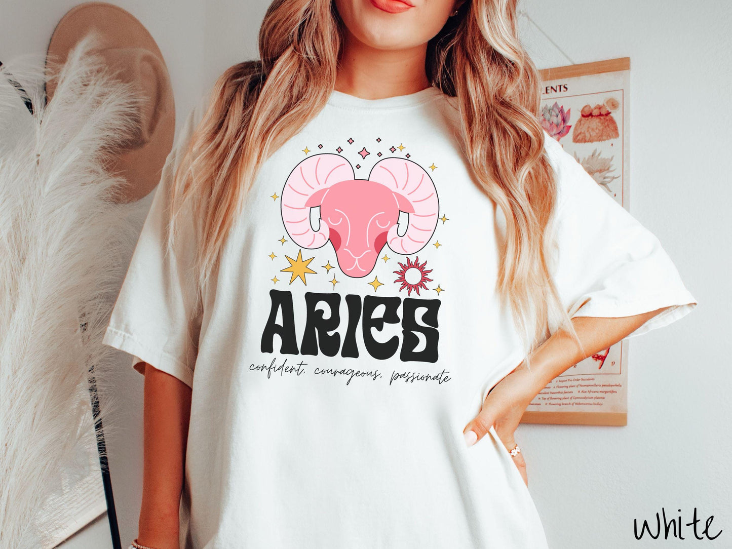 A vintage white colored Comfort Colors T-shirt features a large red ram head surorunded by stars and the sun with the text Aries in large bold font and below that ar the words confident, courageous passionate in cursive font.