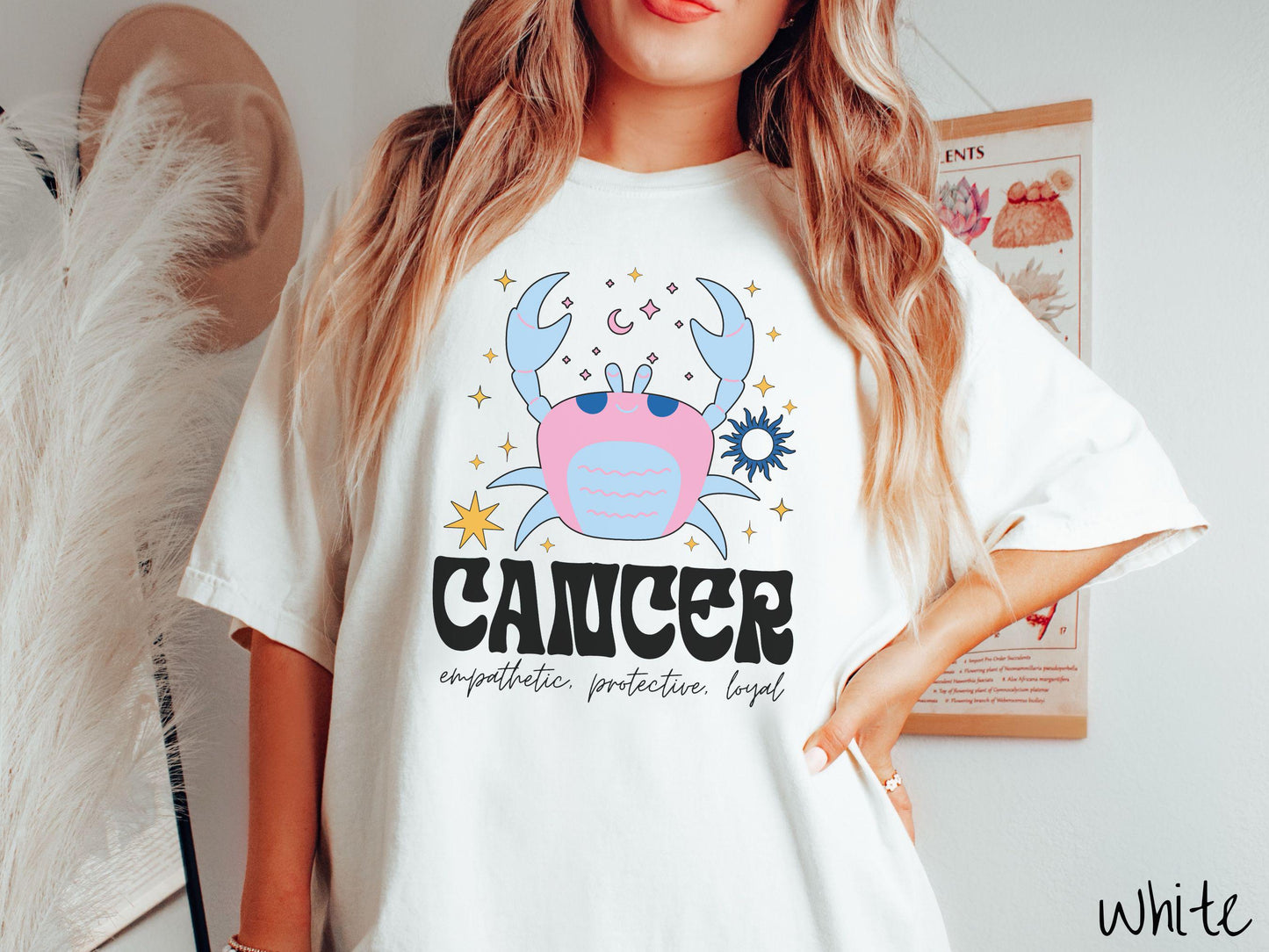 A vintage white colored Comfort Colors T-shirt features a large blue and pink crab surorunded by stars and the sun with the text Cancer in large bold font and below that ar the words empathetic, protective, and loyal in cursive font.