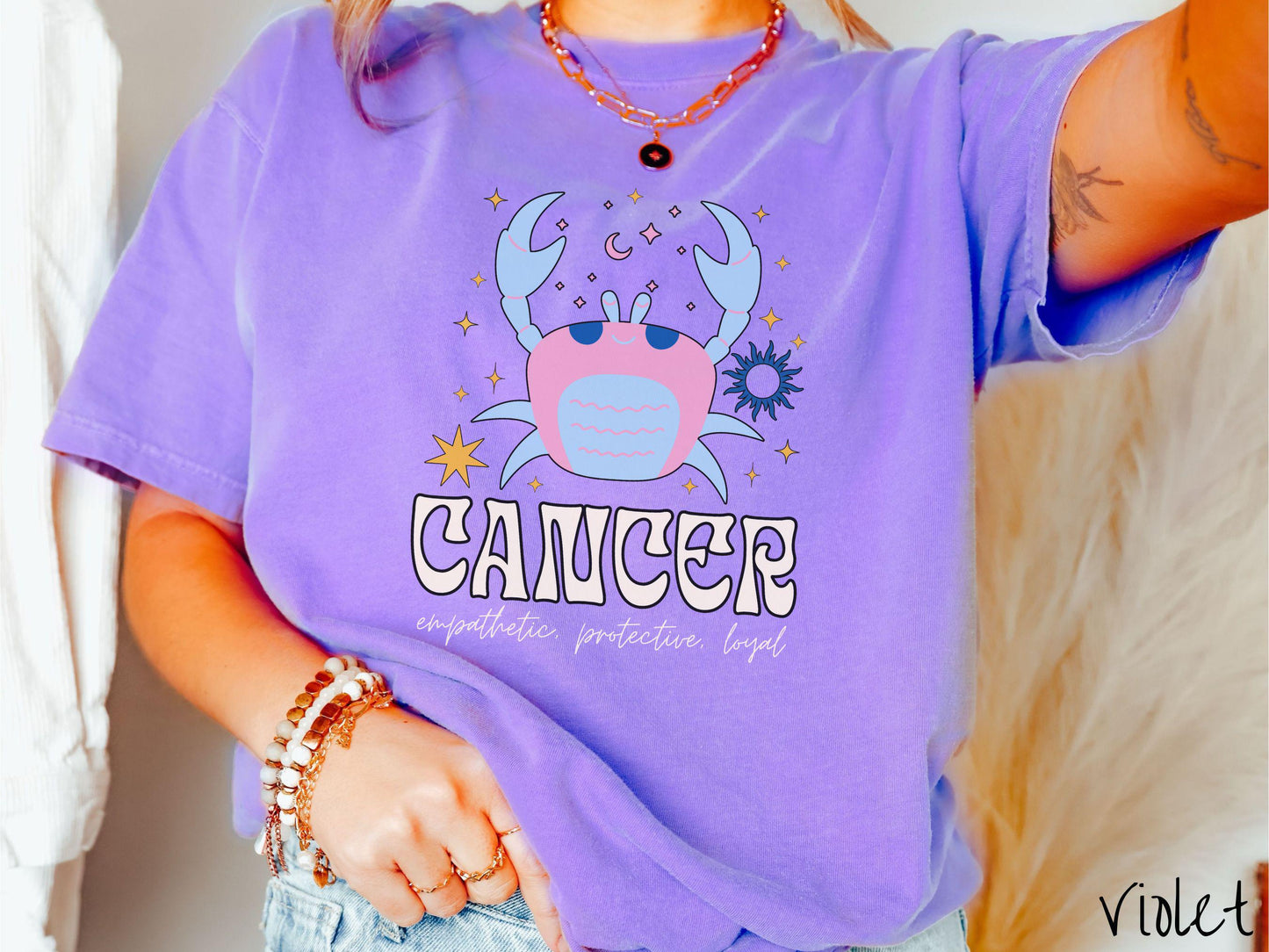 A vintage violet colored Comfort Colors T-shirt features a large blue and pink crab surorunded by stars and the sun with the text Cancer in large bold font and below that ar the words empathetic, protective, and loyal in cursive font.