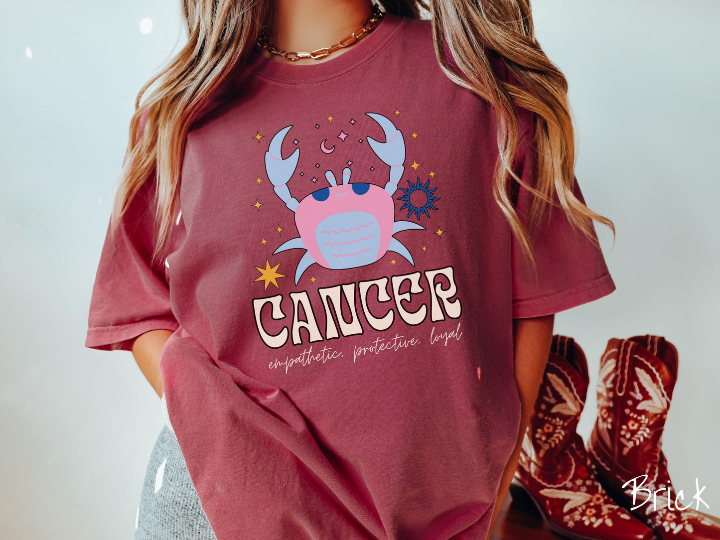 A vintage brick colored Comfort Colors T-shirt features a large blue and pink crab surorunded by stars and the sun with the text Cancer in large bold font and below that ar the words empathetic, protective, and loyal in cursive font.
