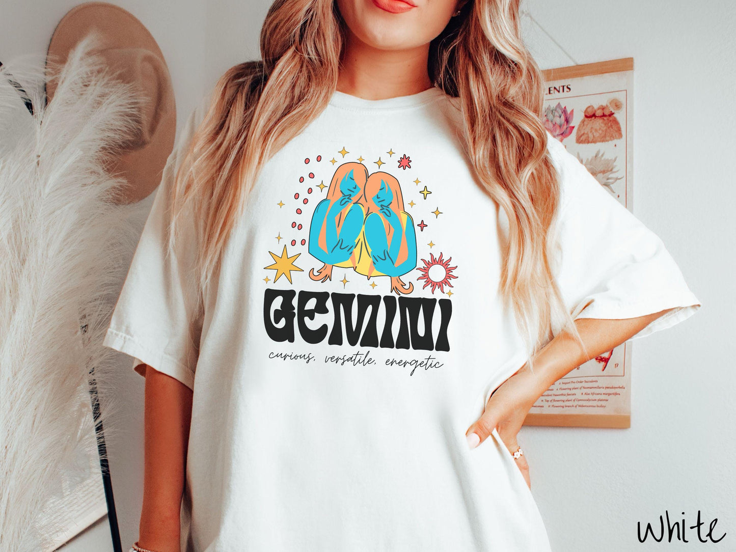 A vintage white colored Comfort Colors T-shirt features the twins Castor and Pollux in blue and orange colors surrounded by stars and the sun with text Gemini in large bold font and below are the words curious, versatile, energetic in cursive font.