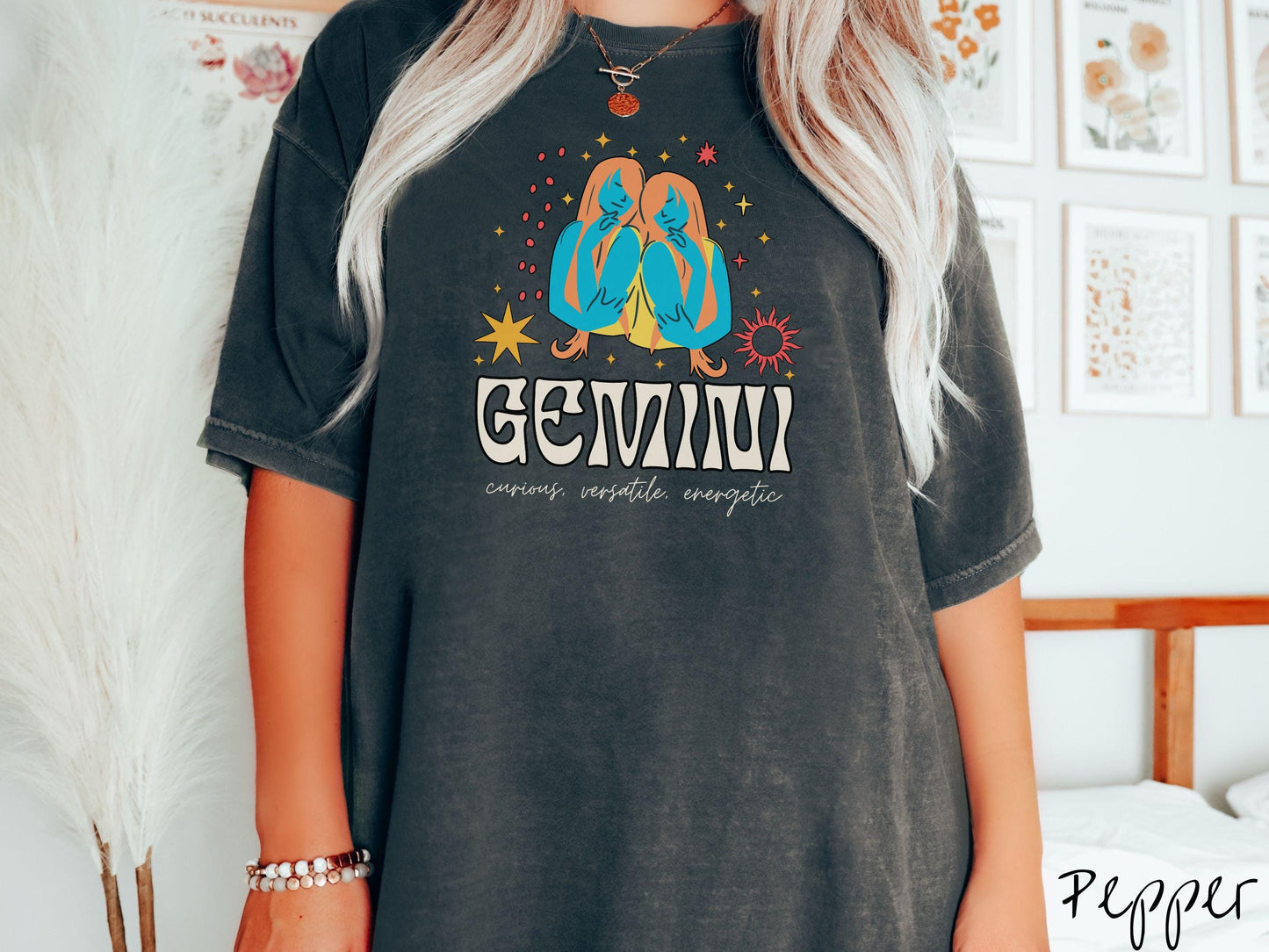 A vintage pepper colored Comfort Colors T-shirt features the twins Castor and Pollux in blue and orange colors surrounded by stars and the sun with text Gemini in large bold font and below are the words curious, versatile, energetic in cursive font.