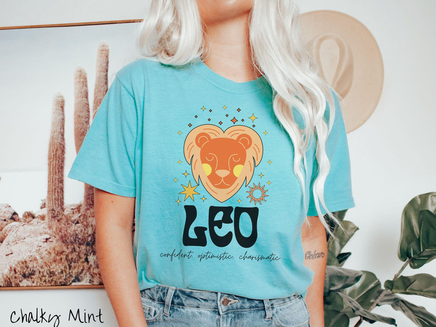 This vintage chalky mint colored Comfort Colors T-shirt features a large orange and yellow lion surrounded by stars and the sun with the text Leo in large bold font and below that are the words confident, optimistic, charismatic in cursive font.