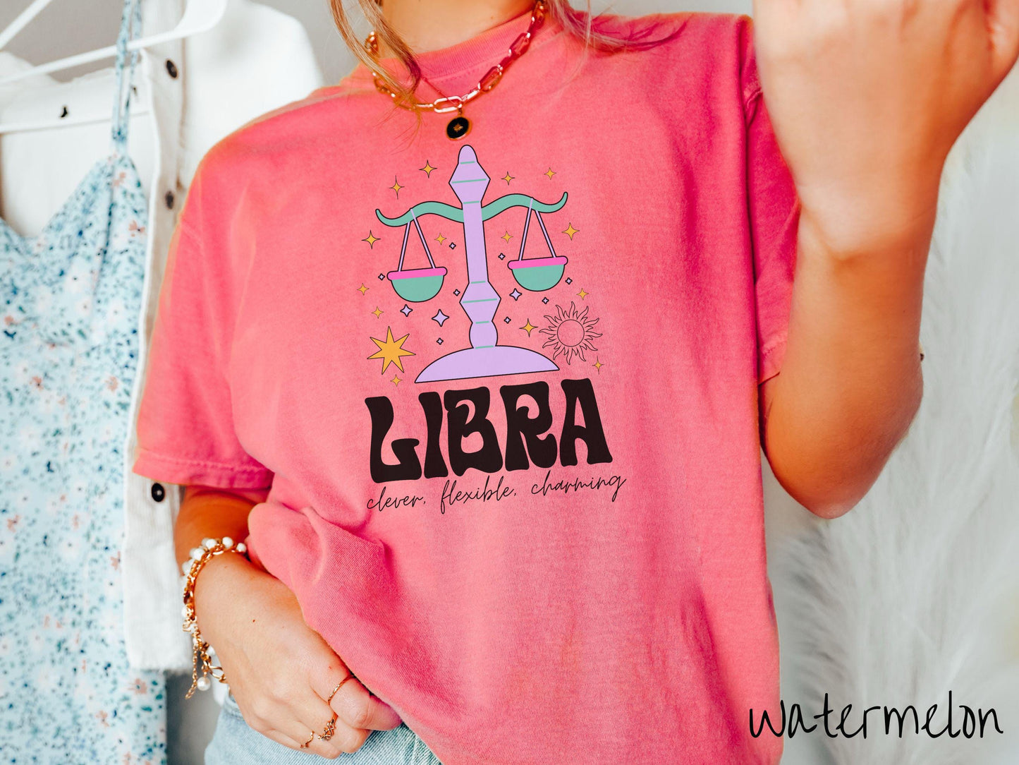A vintage watermelon colored Comfort Colors T-shirt features a large purple and green weight scale surrounded by stars and the sun with the text Libra in large bold font and below that are the words clever, flexible, charming in cursive font.