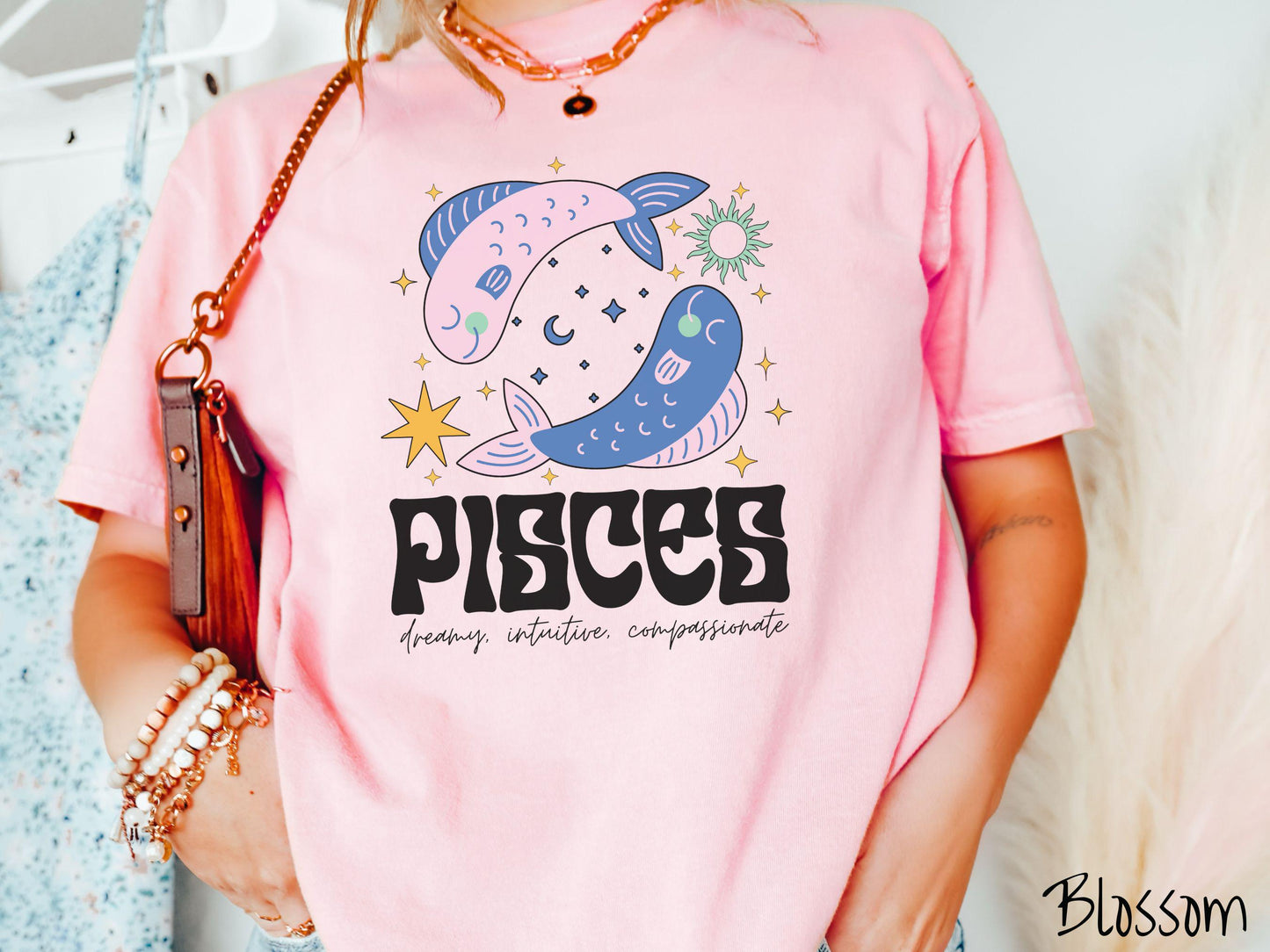 A vintage blossom colored Comfort Colors T-shirt features two large pink and blue fish surrounded by stars and the sun with the text Pisces in large bold font and below that are the words dreamy, intuitive, compassionate in cursive font.