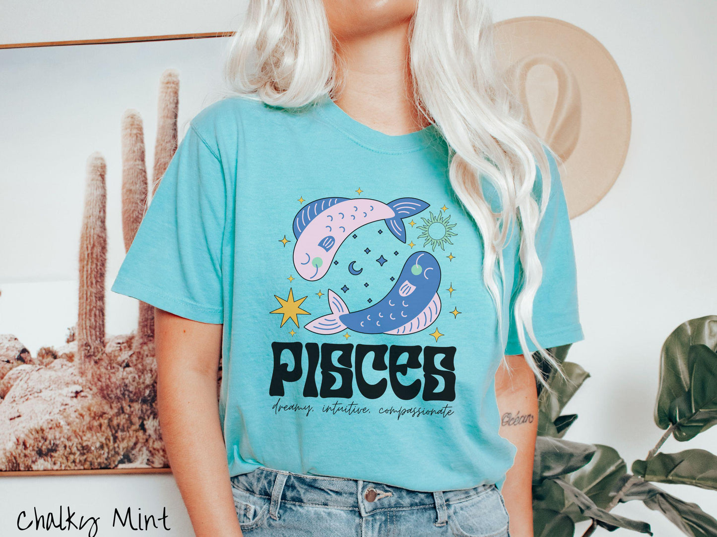 A vintage chalky mint colored Comfort Colors T-shirt features two large pink and blue fish surrounded by stars and the sun with the text Pisces in large bold font and below that are the words dreamy, intuitive, compassionate in cursive font.