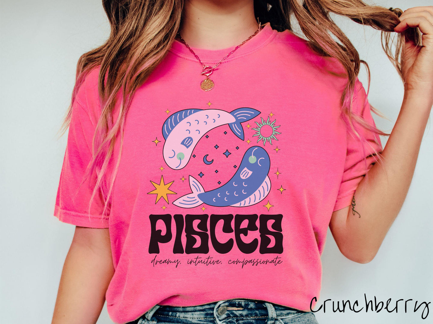A vintage crunchberry colored Comfort Colors T-shirt features two large pink and blue fish surrounded by stars and the sun with the text Pisces in large bold font and below that are the words dreamy, intuitive, compassionate in cursive font.