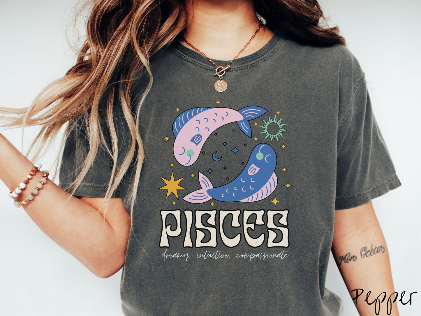 A vintage pepper colored Comfort Colors T-shirt features two large pink and blue fish surrounded by stars and the sun with the text Pisces in large bold font and below that are the words dreamy, intuitive, compassionate in cursive font.