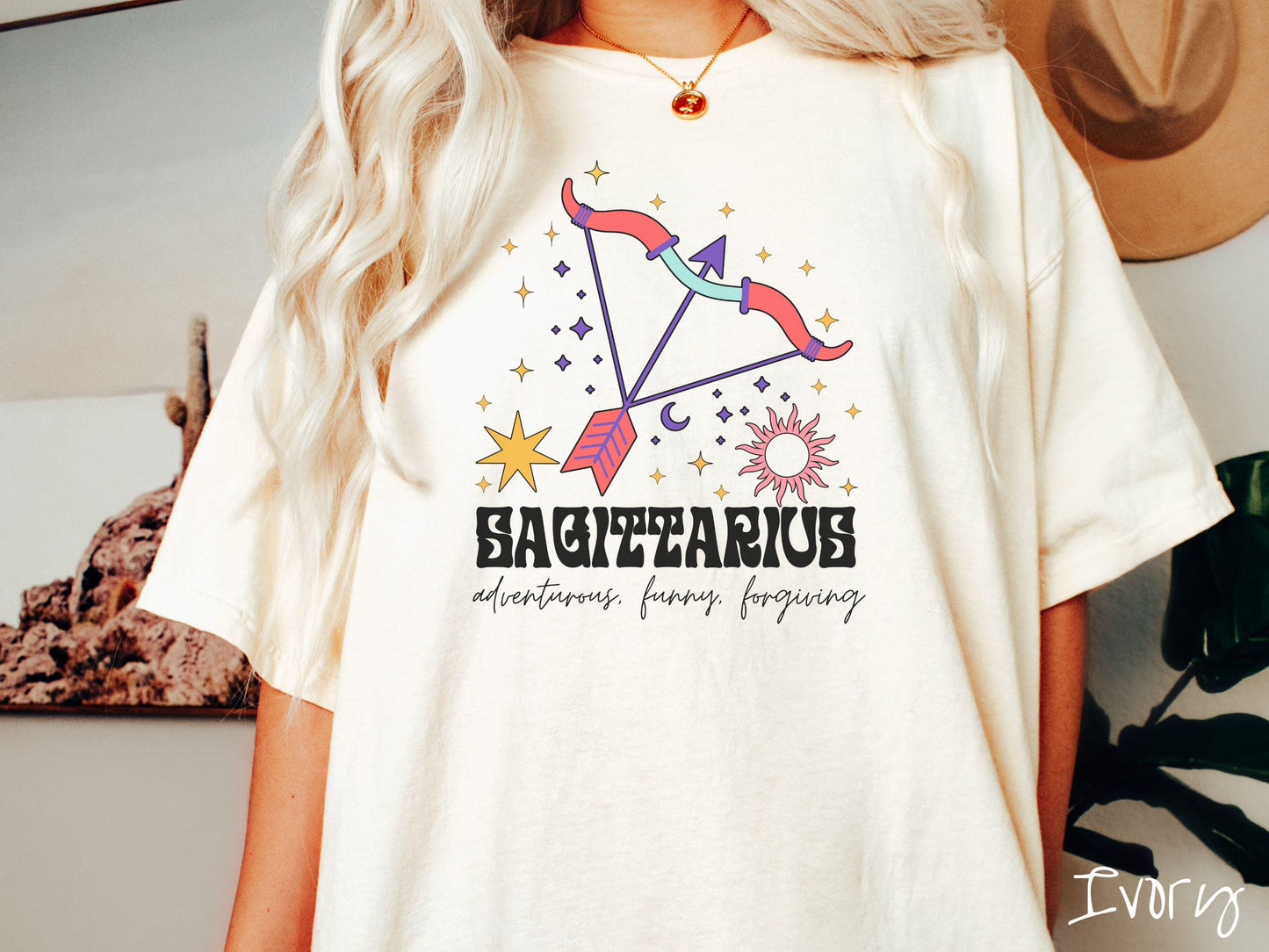 A vintage ivory colored Comfort Colors T-shirt features a large red and purple bow and arrow surrounded by stars and the sun with the text Sagittarius in large bold font and below that are the words adventurous, funny, forgiving in cursive font.