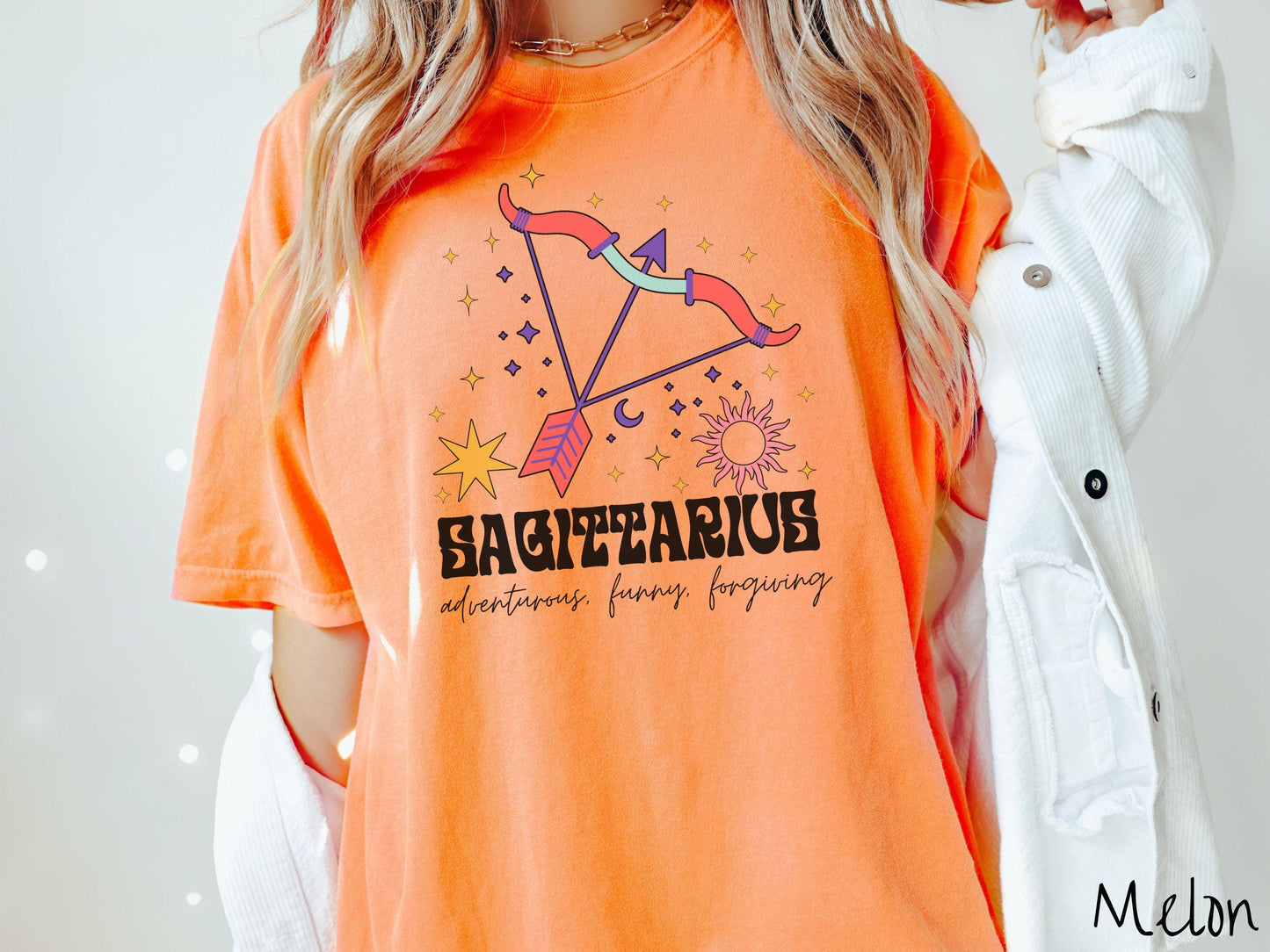 A vintage melon colored Comfort Colors T-shirt features a large red and purple bow and arrow surrounded by stars and the sun with the text Sagittarius in large bold font and below that are the words adventurous, funny, forgiving in cursive font.