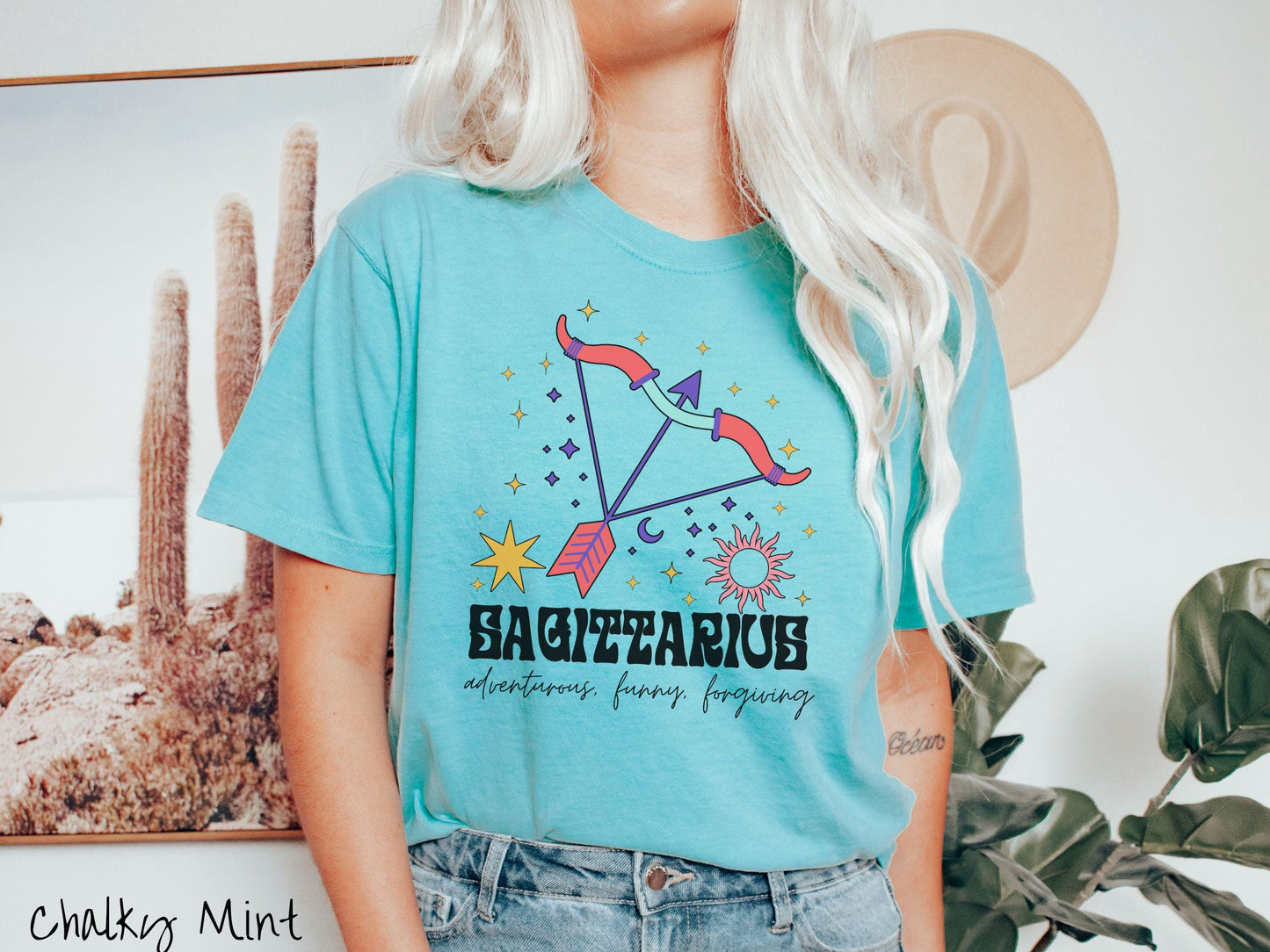 A vintage chalky mint colored Comfort Colors T-shirt features a large red and purple bow and arrow surrounded by stars and sun with the text Sagittarius in large bold font and below that are the words adventurous, funny, forgiving in cursive font.