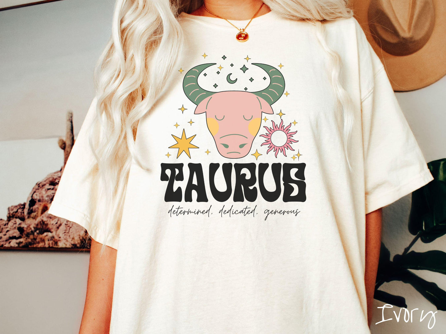 A vintage ivory colored Comfort Colors T-shirt features a large pink and green bull surrounded by stars and the sun with the text Taurus in large bold font and below that are the words determined, dedicated, generous in cursive font.