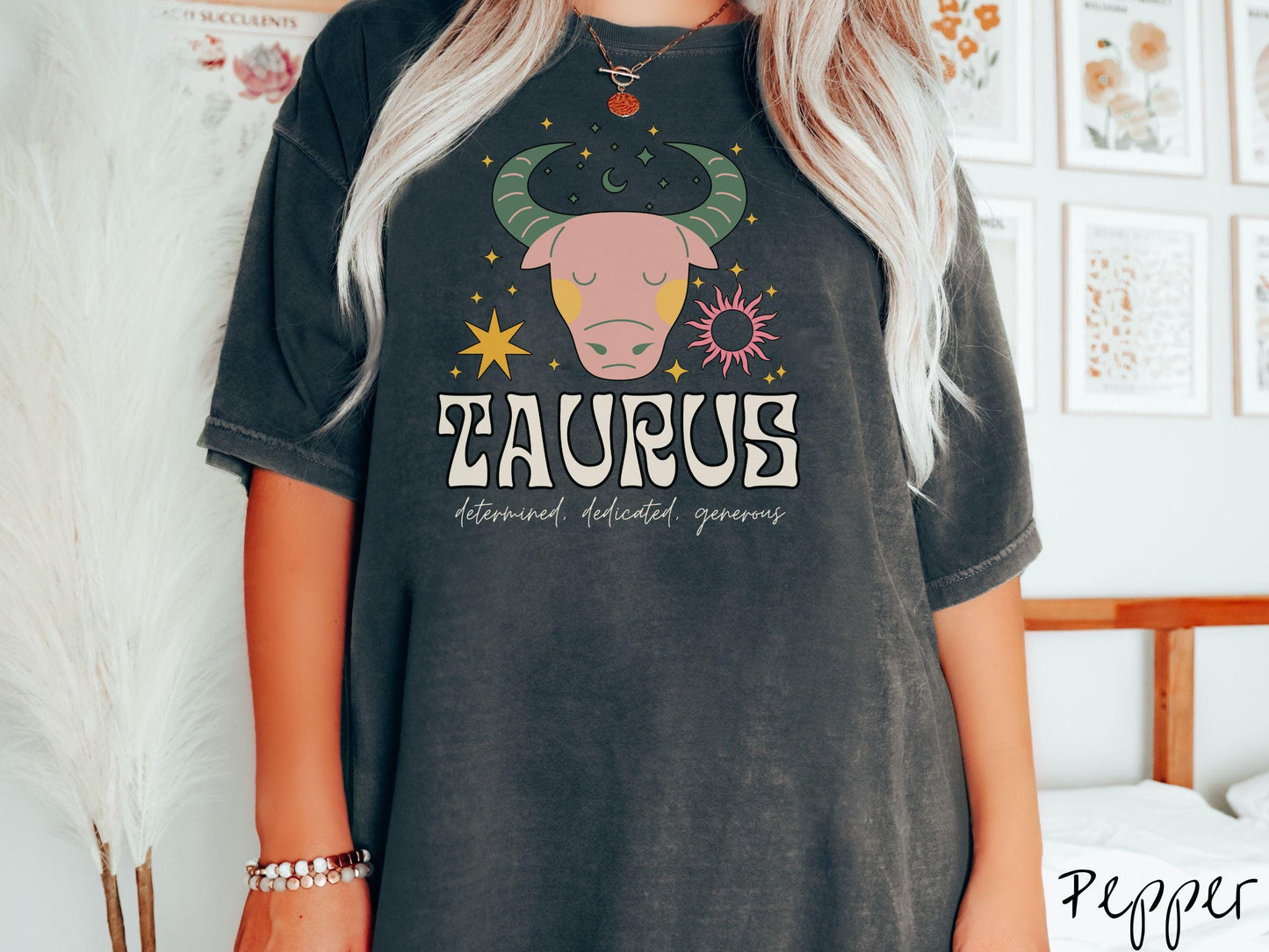 A vintage pepper colored Comfort Colors T-shirt features a large pink and green bull surrounded by stars and the sun with the text Taurus in large bold font and below that are the words determined, dedicated, generous in cursive font.