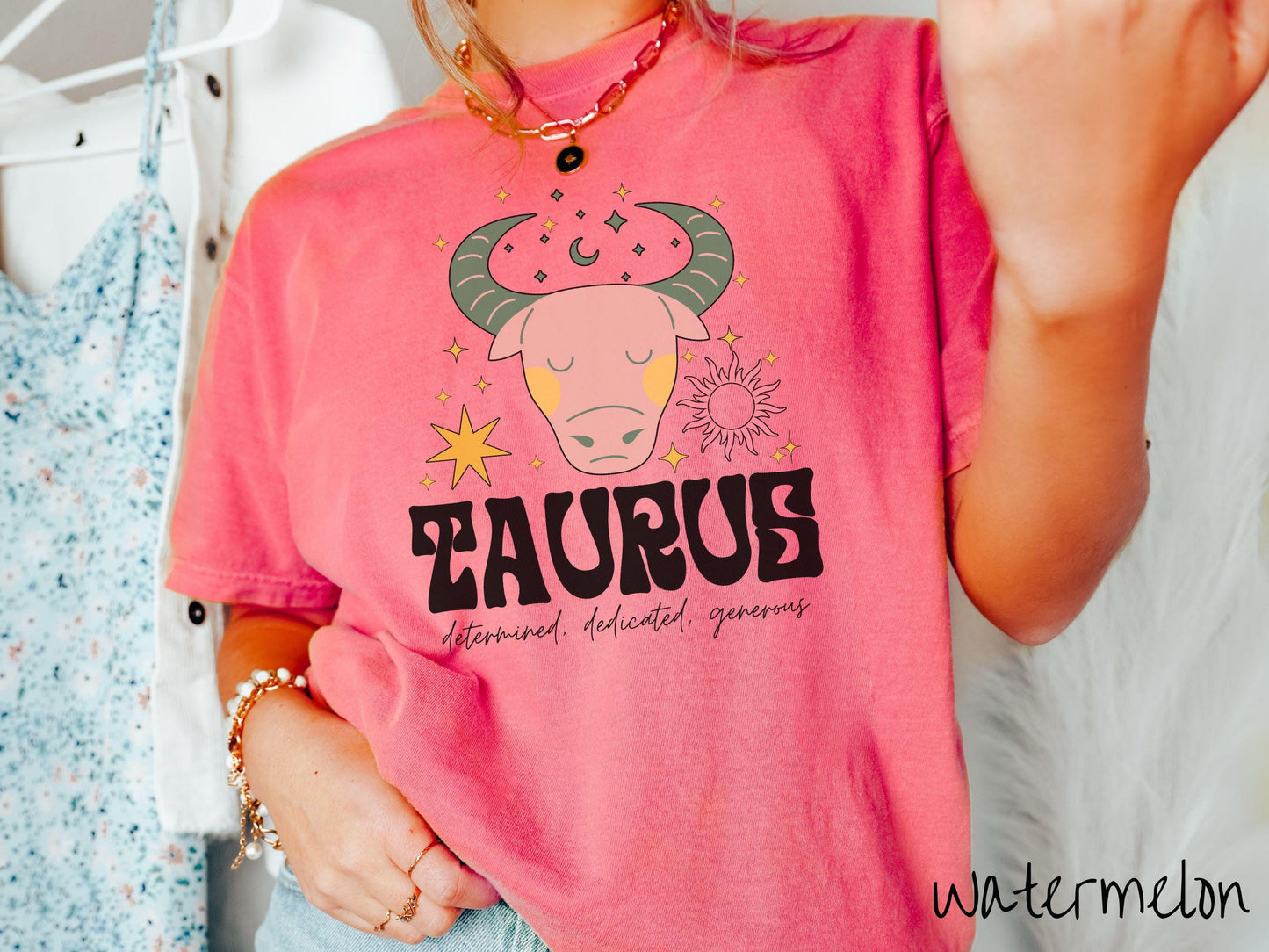 A vintage watermelon colored Comfort Colors T-shirt features a large pink and green bull surrounded by stars and the sun with the text Taurus in large bold font and below that are the words determined, dedicated, generous in cursive font.