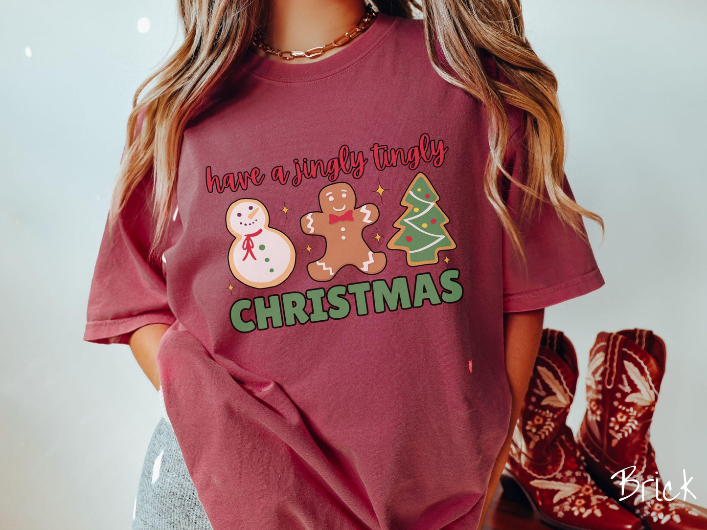 A vintage brick colored Christmas Comfort Colors t-shirt featuring a snowman iced cookie, a gingerbread man cookie, and a Christmas tree cookie. The text reads 'have a jingly tingly Christmas' in red and green font.