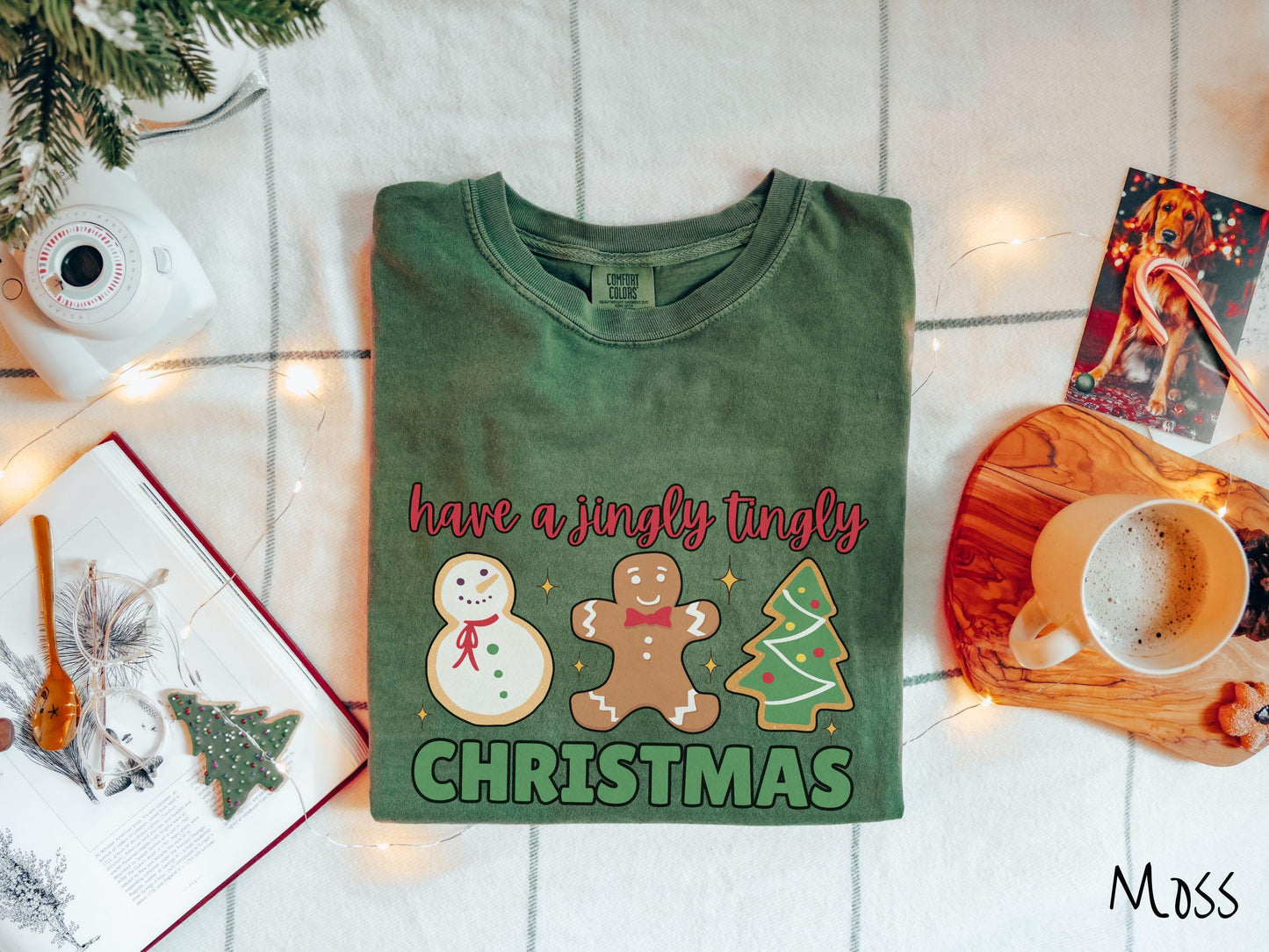 A vintage moss colored Christmas Comfort Colors t-shirt featuring a snowman iced cookie, a gingerbread man cookie, and a Christmas tree cookie. The text reads 'have a jingly tingly Christmas' in red and green font.