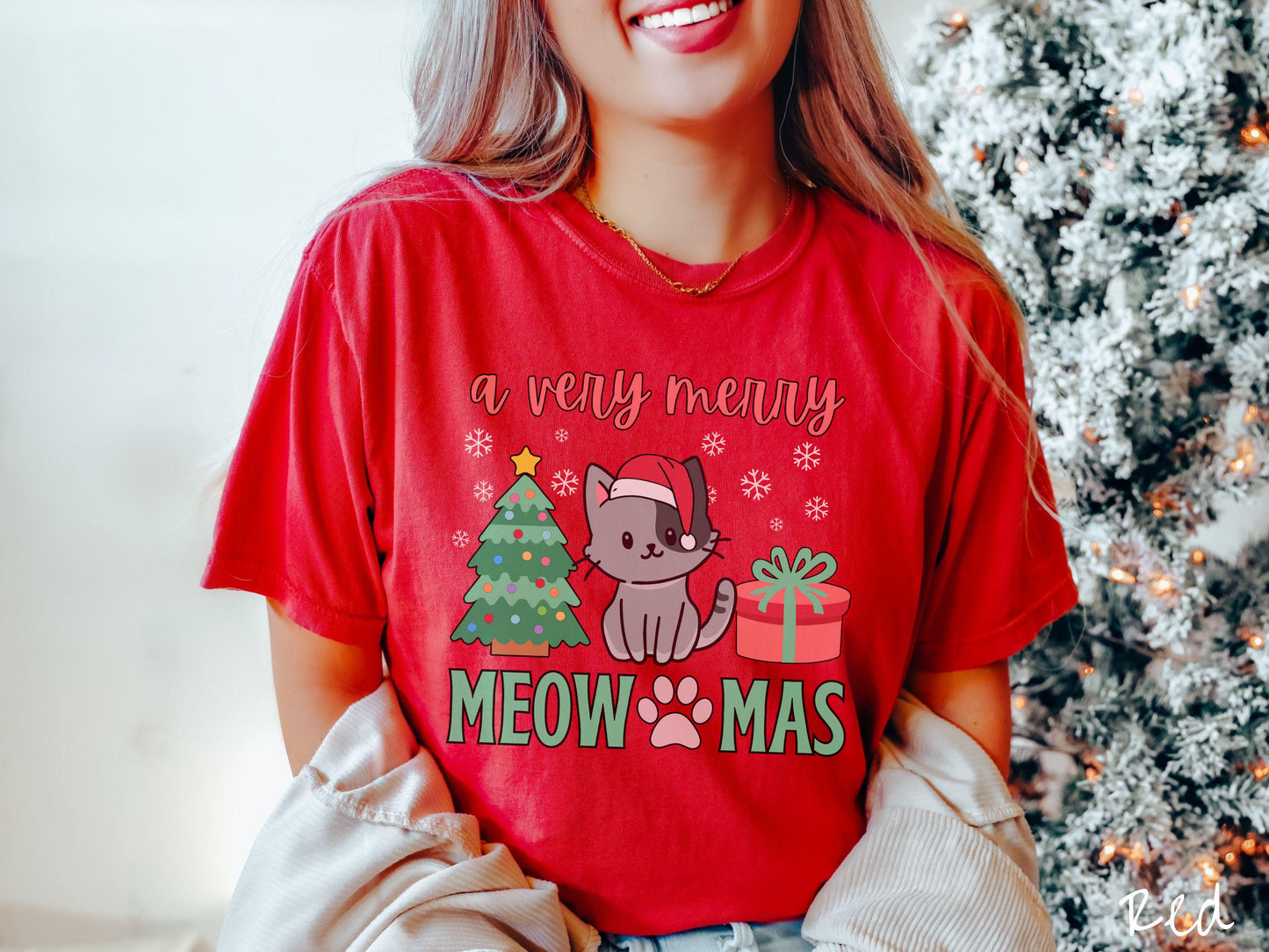 A vintage red colored Comfort Colors T-shirt with the text 'a very merry Christmas' in red and green font, featuring a grey cat wearing a red Santa hat sitting between a Christmas tree and a present with white snowflakes hanging in the air.