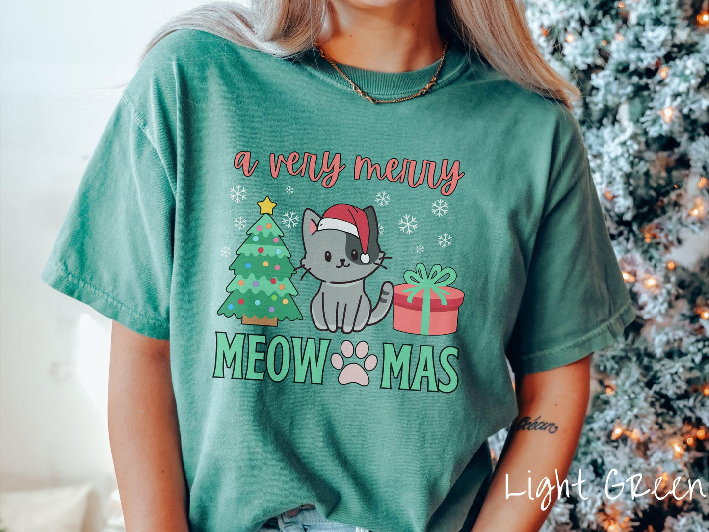 A vintage light green colored Comfort Colors T-shirt with the text 'a very merry Christmas' in red and green font, featuring a grey cat wearing a red Santa hat sitting between a Christmas tree and a present with white snowflakes hanging in the air.