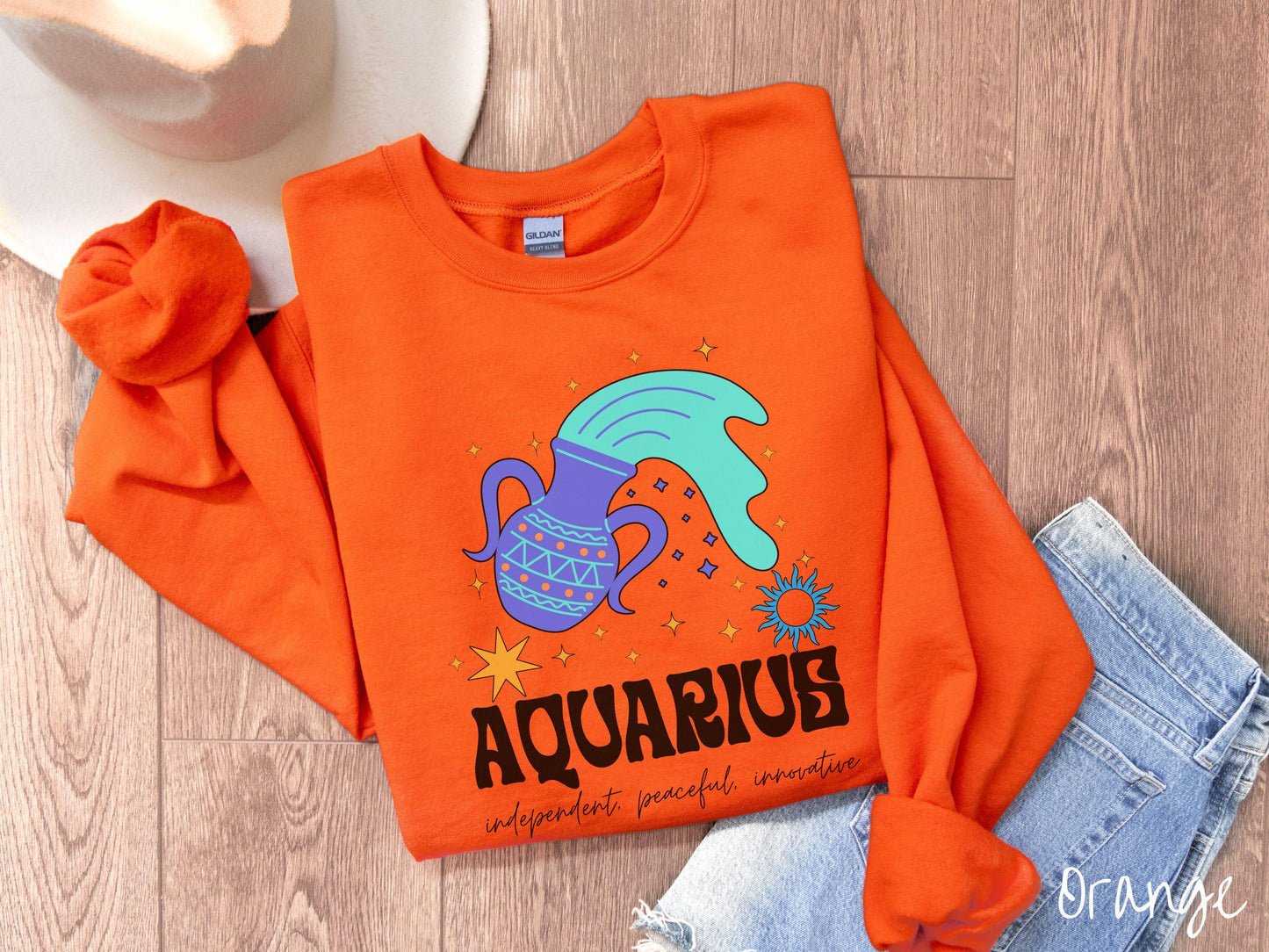 This orange colored vintage Comfort Colors sweatshirt features a large purple urn with blue water flowing out of it, surrounded by stars, with the text Aquarius and the words independent, peaceful, innovative.