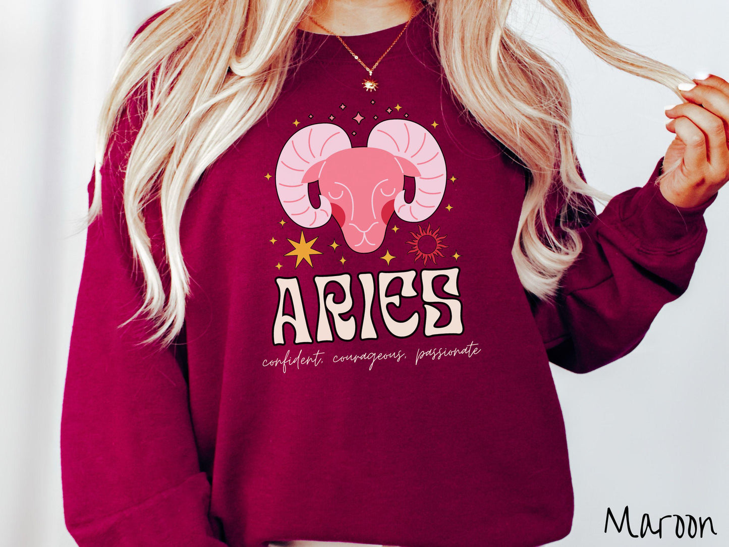 A vintage maroon colored Comfort Colors sweatshirt features a large red ram head surrounded by stars and the sun, with the text Aries in large bold font and below that are the words confident, courageous, passionate in cursive font.