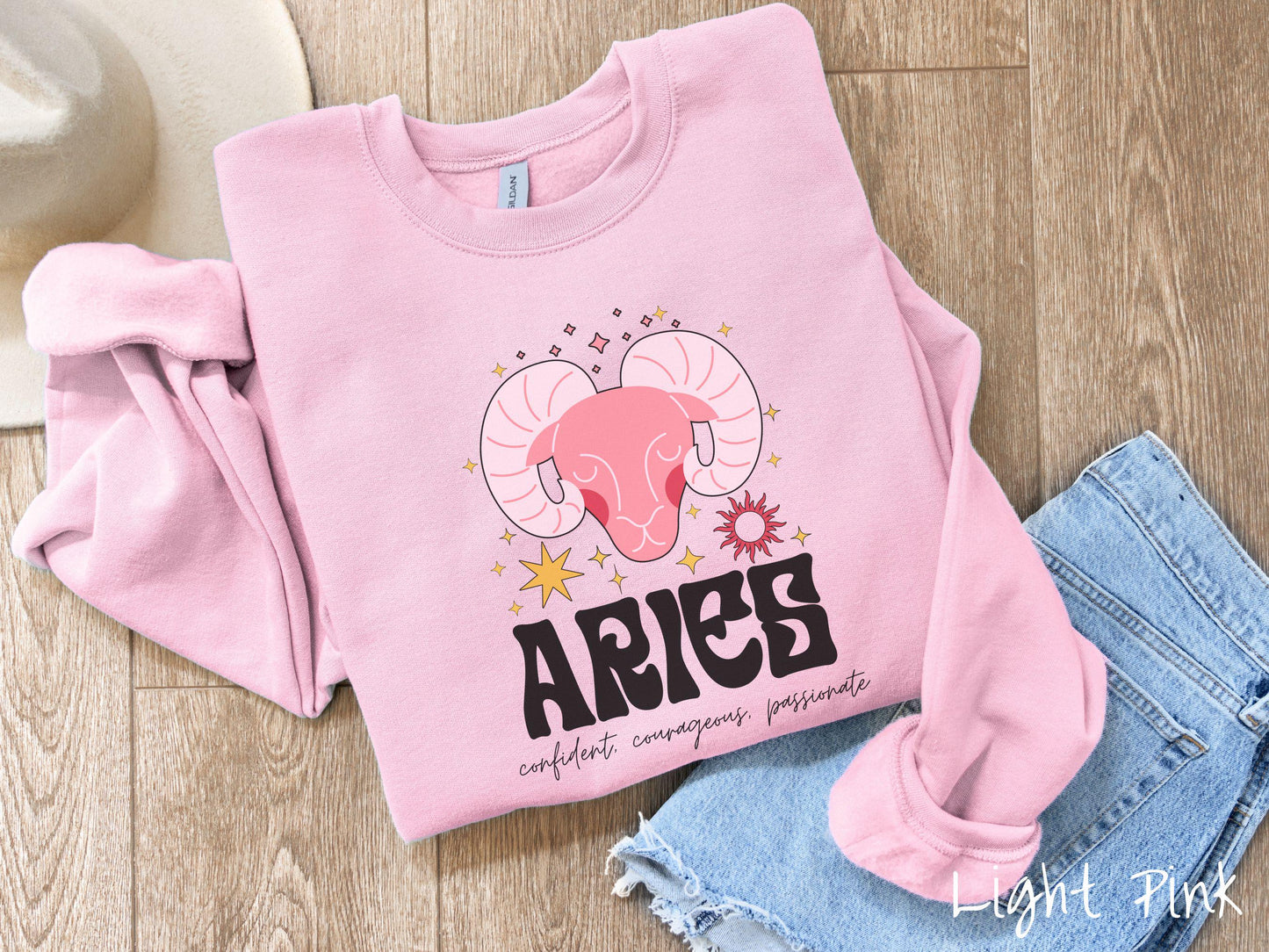 A vintage light pink colored Comfort Colors sweatshirt features a large red ram head surrounded by stars and the sun, with the text Aries in large bold font and below that are the words confident, courageous, passionate in cursive font.