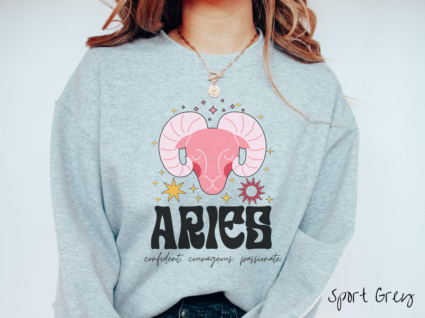 A vintage sport grey colored Comfort Colors sweatshirt features a large red ram head surrounded by stars and the sun, with the text Aries in large bold font and below that are the words confident, courageous, passionate in cursive font.