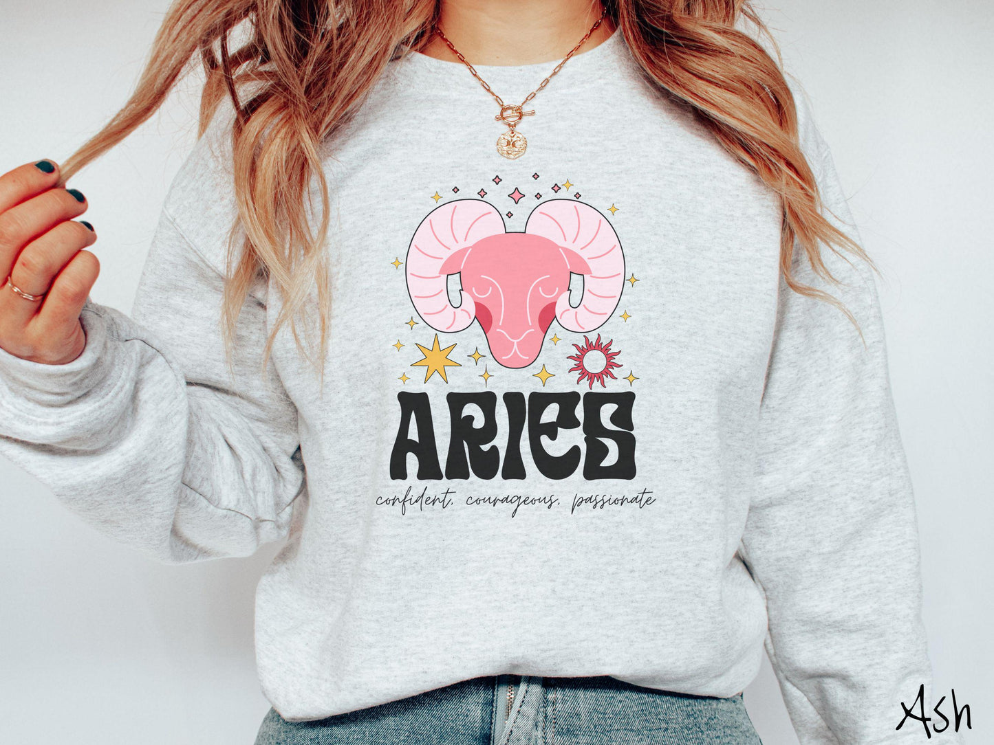 A vintage ash colored Comfort Colors sweatshirt features a large red ram head surrounded by stars and the sun, with the text Aries in large bold font and below that are the words confident, courageous, passionate in cursive font.