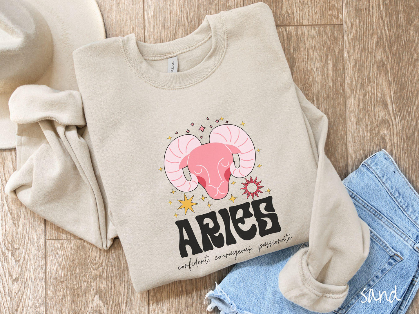 A vintage sand colored Comfort Colors sweatshirt features a large red ram head surrounded by stars and the sun, with the text Aries in large bold font and below that are the words confident, courageous, passionate in cursive font.