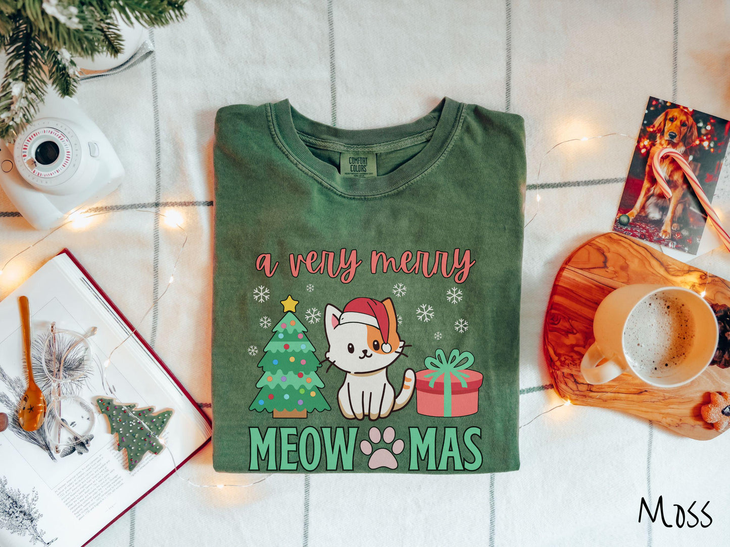 A vintage moss colored Comfort Colors T-shirt with the text a very merry Christmas in red and green font, featuring a grey cat wearing a red Santa hat sitting between a Christmas tree and a present with white snowflakes hanging in the air.
