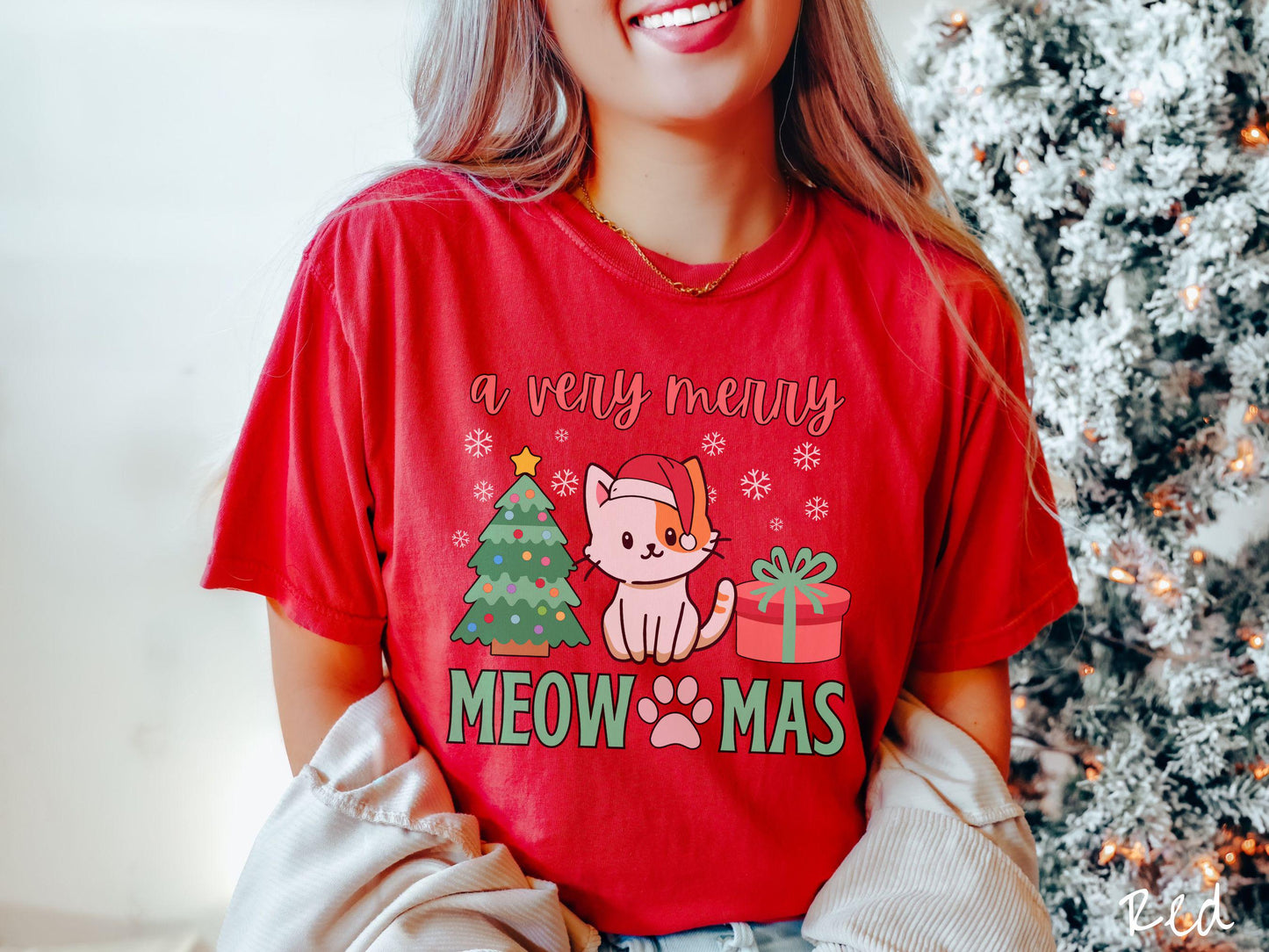 A vintage red colored Comfort Colors T-shirt with the text a very merry Christmas in red and green font, featuring a grey cat wearing a red Santa hat sitting between a Christmas tree and a present with white snowflakes hanging in the air.